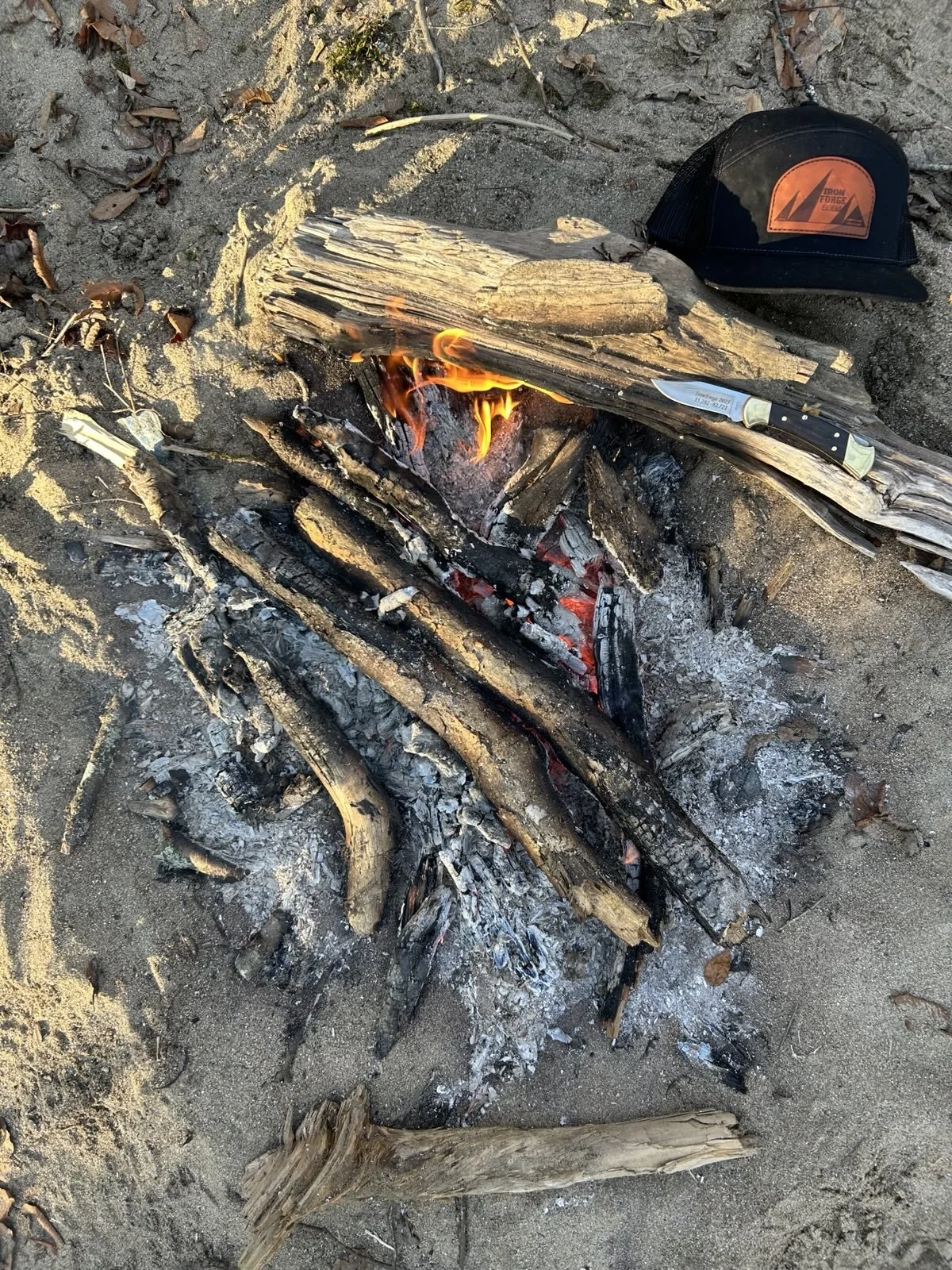 Small campfire on sandy ground with burning logs, a knife resting on a wood piece next to the fire, and a cap nearby.