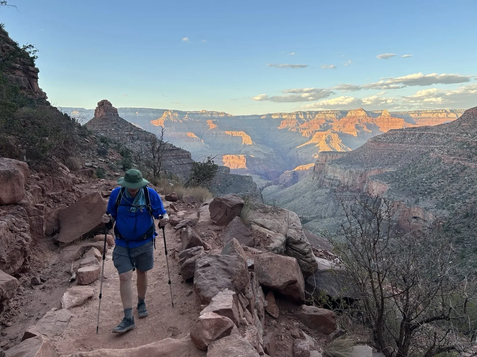 A hiker wearing a blue jacket, gray shorts, and a green hat walking on a rocky trail in the Grand Canyon during daylight, with layered canyon cliffs and a partly cloudy sky in the background.