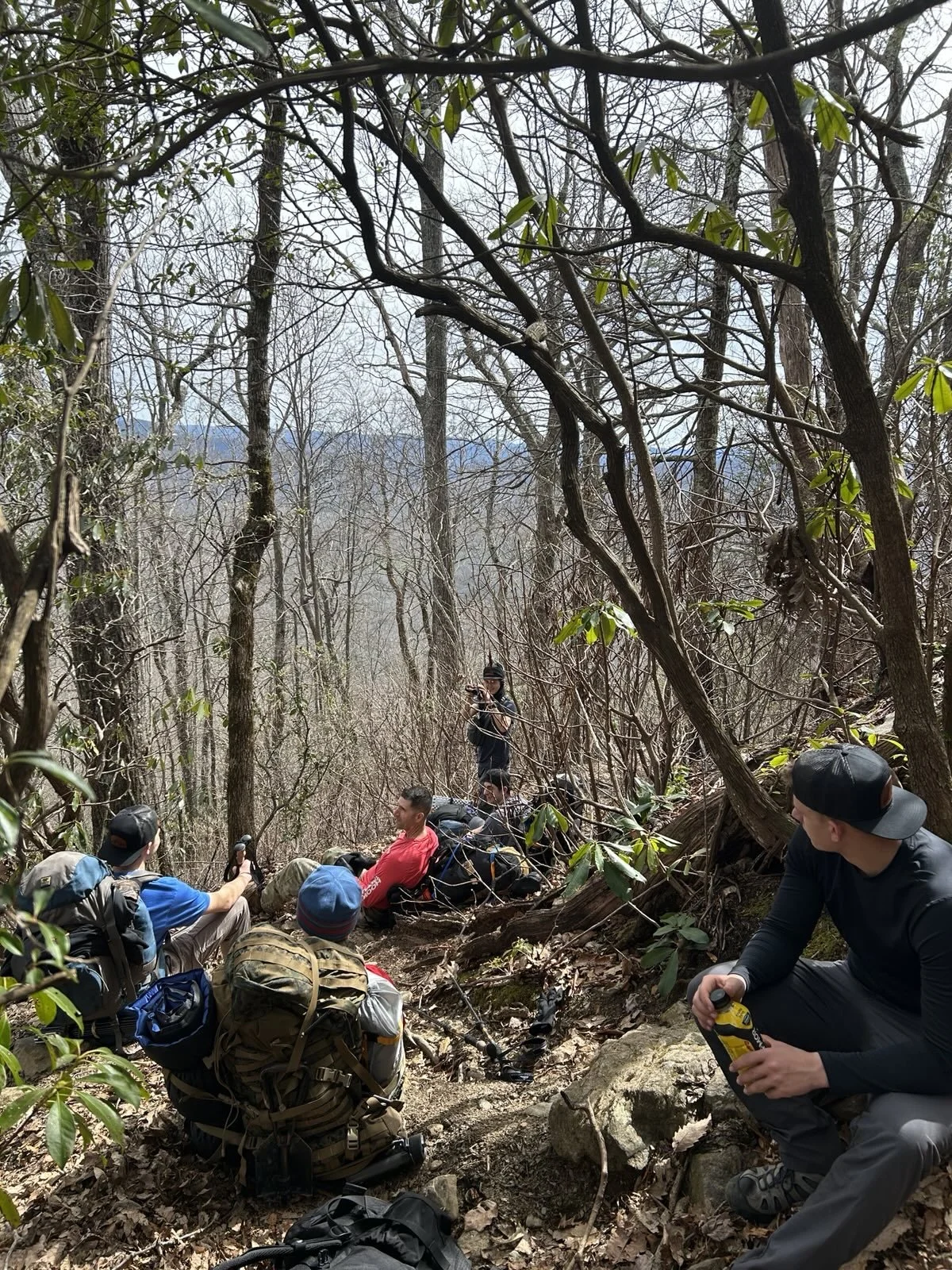 Group of hikers resting on a wooded mountain trail surrounded by leafless trees, some with green leaves, with backpacks and gear, one person taking photos, and others sitting on rocks or the ground.