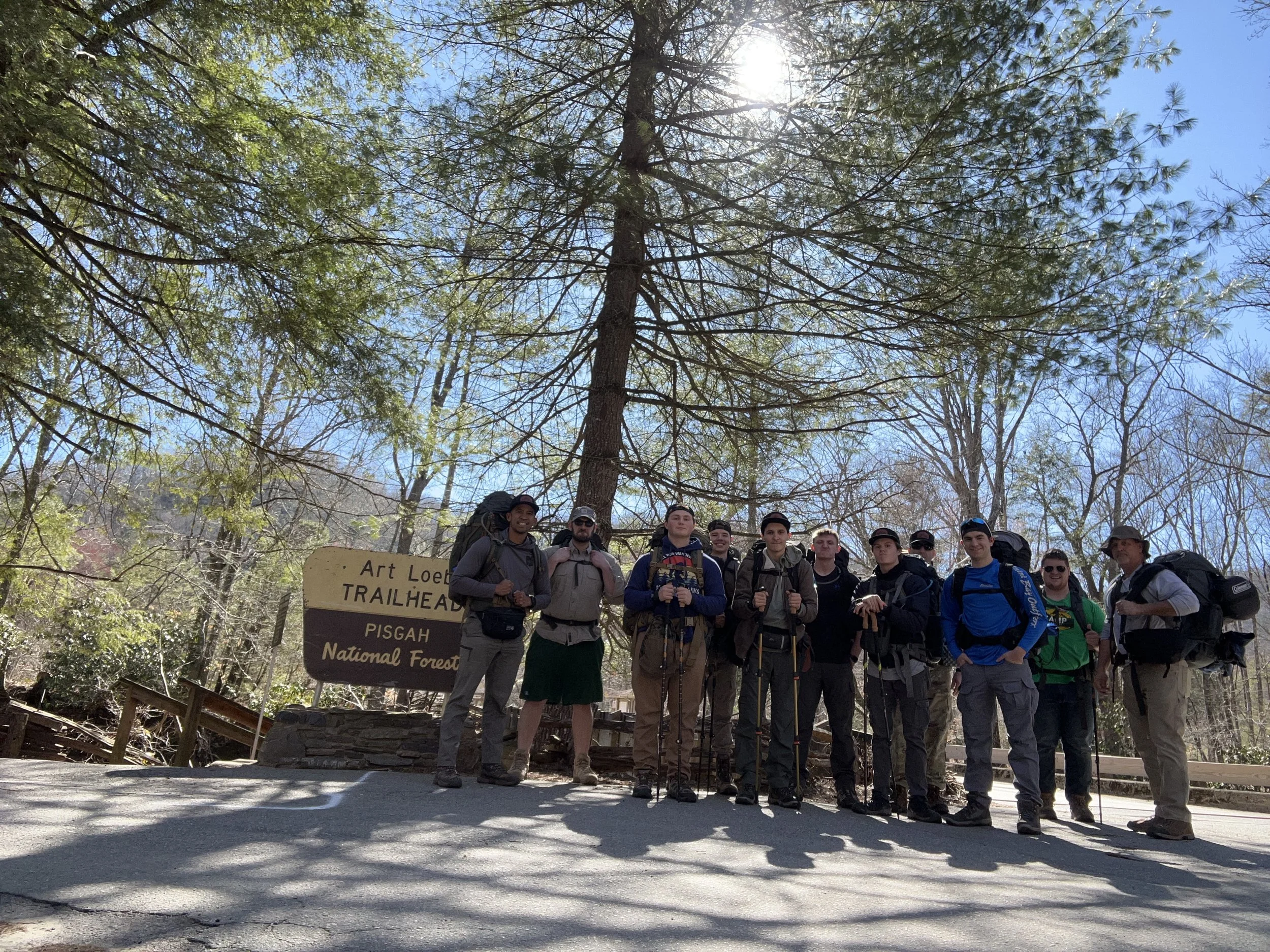 Group of hikers standing in front of a trailhead sign in a forest, with large trees and blue sky visible.