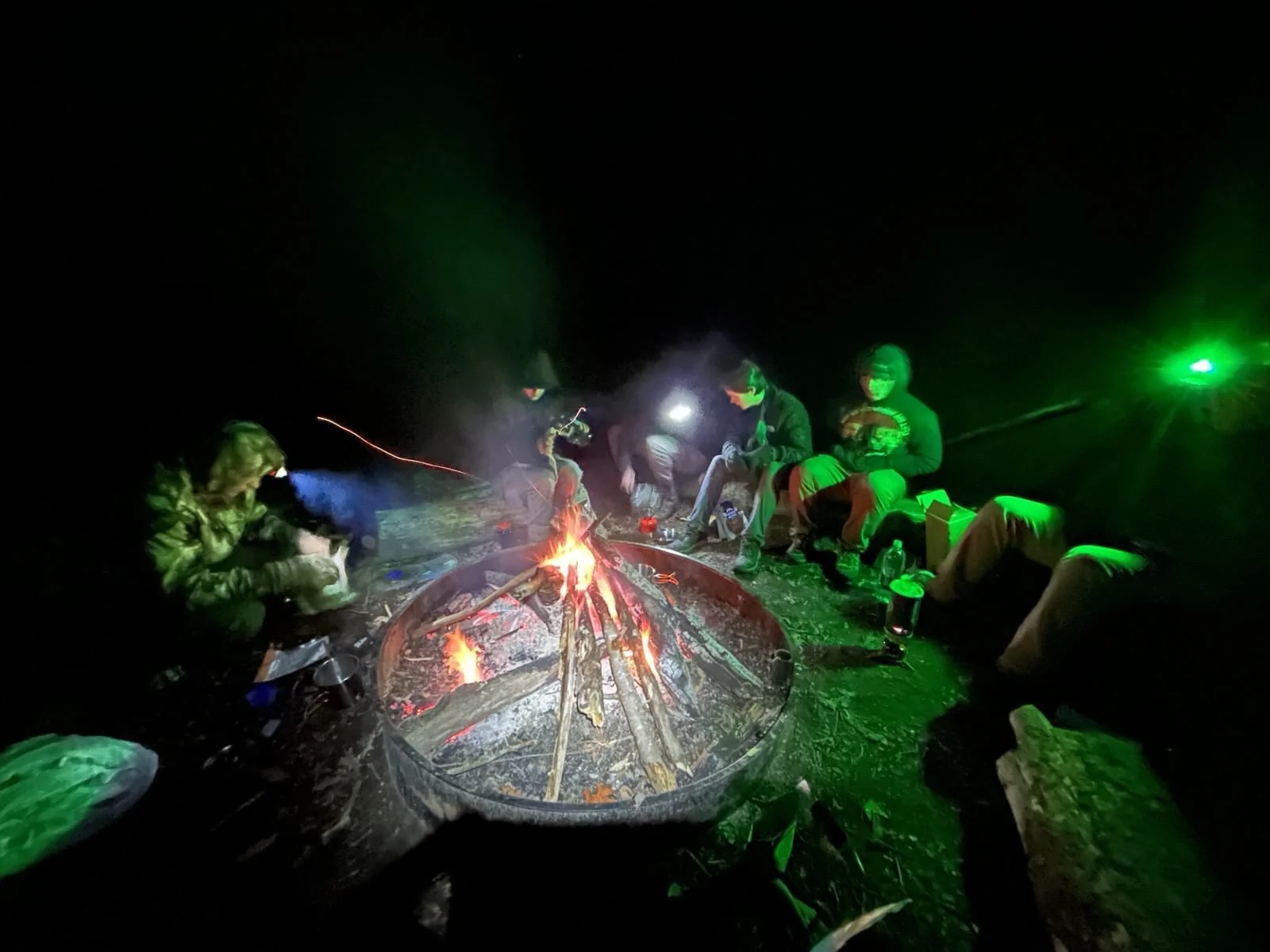People sitting around a campfire at night, wearing jackets and masks, with green and blue lights illuminating the scene.
