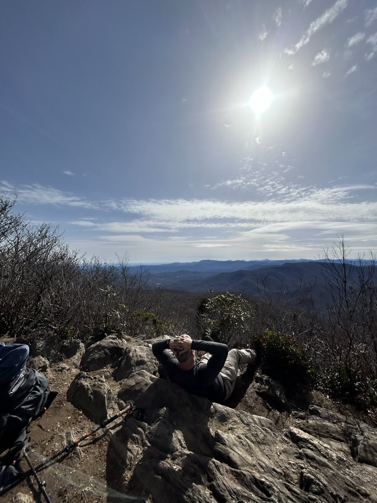 A person relaxing on a rocky mountaintop, overlooking a vast landscape of mountains and a clear sky with the sun shining brightly.