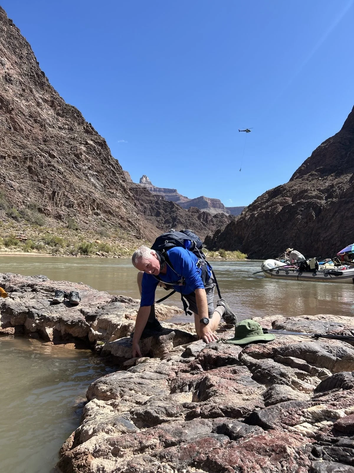 A man in outdoor gear on a rocky riverbank, climbing onto rocks in a canyon with steep cliffs on both sides. A helicopter is flying overhead, towing a container, and there is a boat on the river with people and supplies.