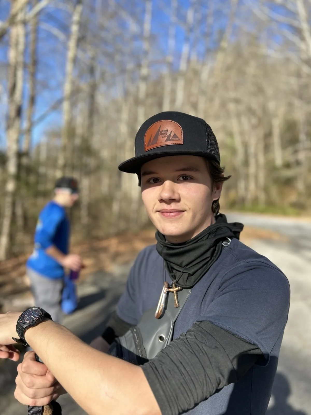 Young man outdoors wearing a hat, watch, and neck gaiter, with another person in the background in a wooded area.