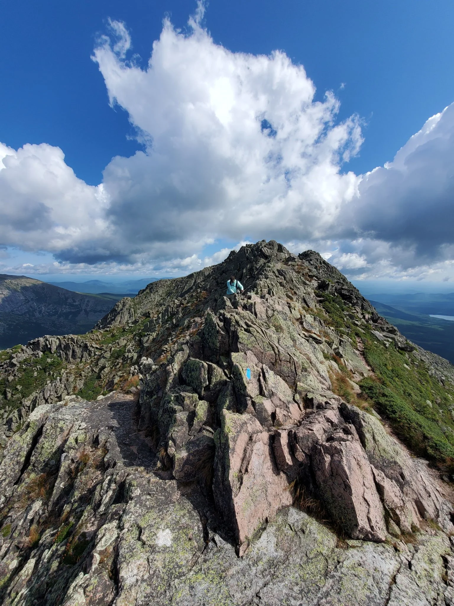 Hiker climbing rocky mountain ridge with cloudy sky and distant landscape in background.