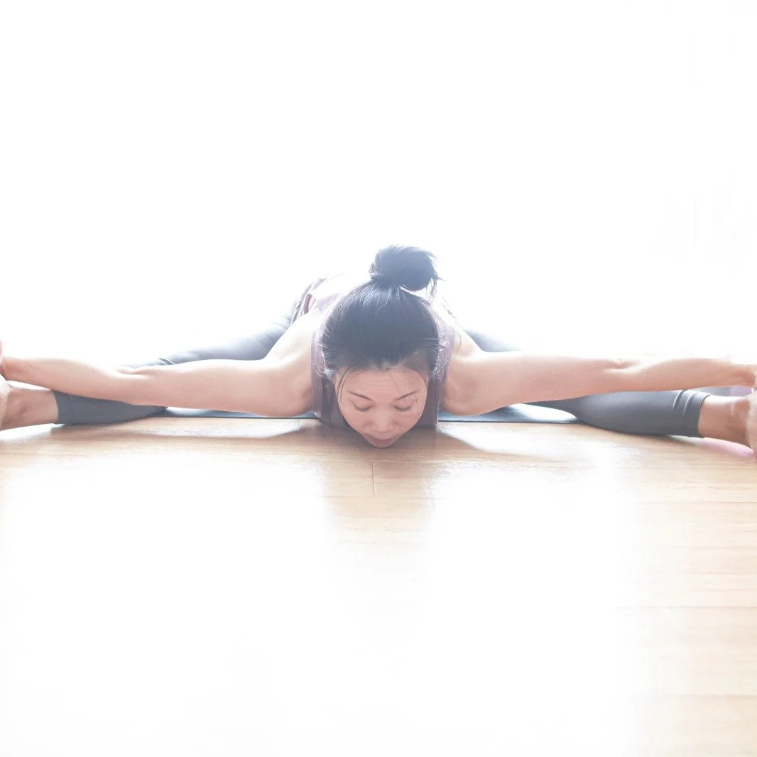 Mary Ma Yoga practicing yoga in a wide straddle split on a wooden floor, performing a forward bend with her head down.
