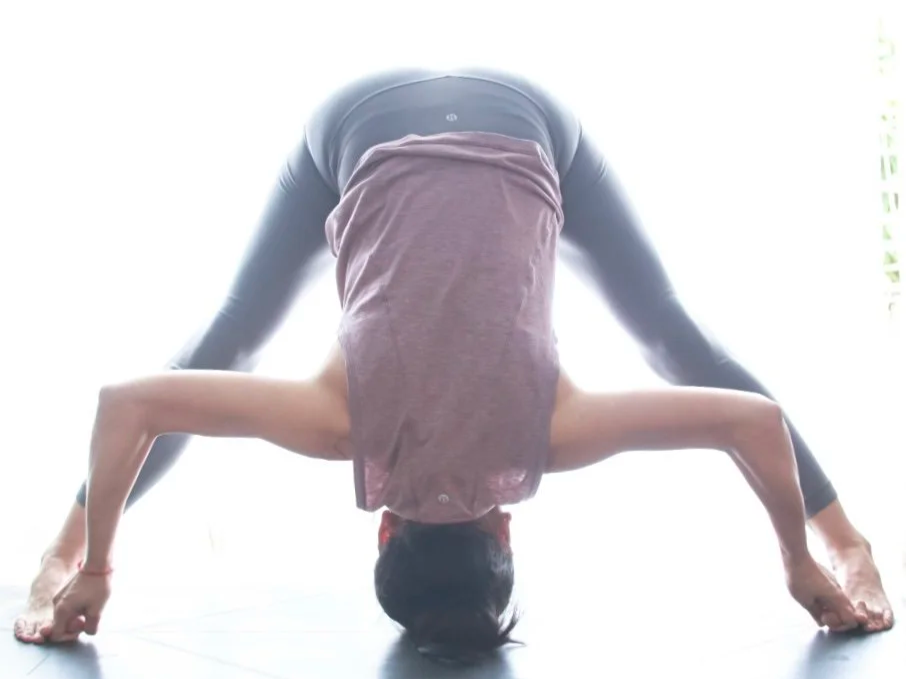 Mary Ma Yoga in downward dog yoga pose on a mat, wearing a pink shirt and black leggings, with a bright light background.