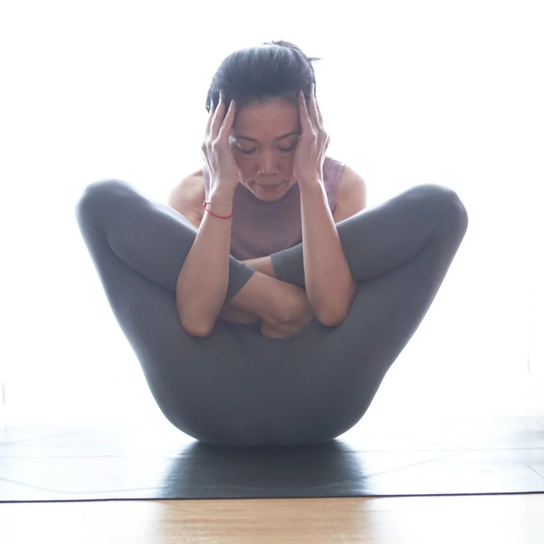 Mary Ma Yoga practicing yoga in a challenging pose with her legs crossed and her head resting in her hands.