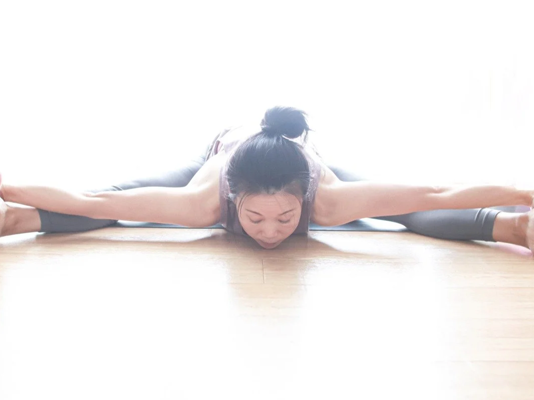 Mary Ma Yoga doing a full split yoga pose on the floor with bright light behind.
