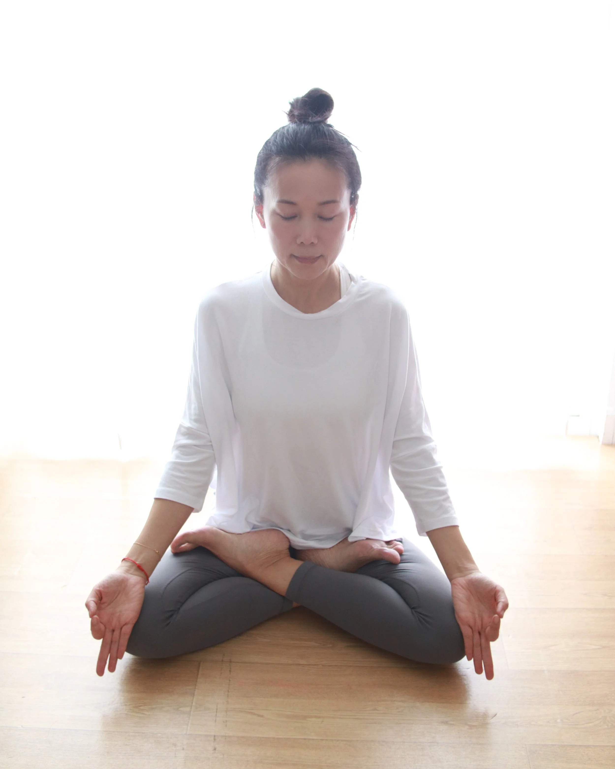 Mary Ma Yoga practicing yoga in a seated meditation pose on a wooden floor, with binding her left foot with her right arm, and her right foot with her left arm, sitting cross-legged with eyes closed and hands resting on her knees.