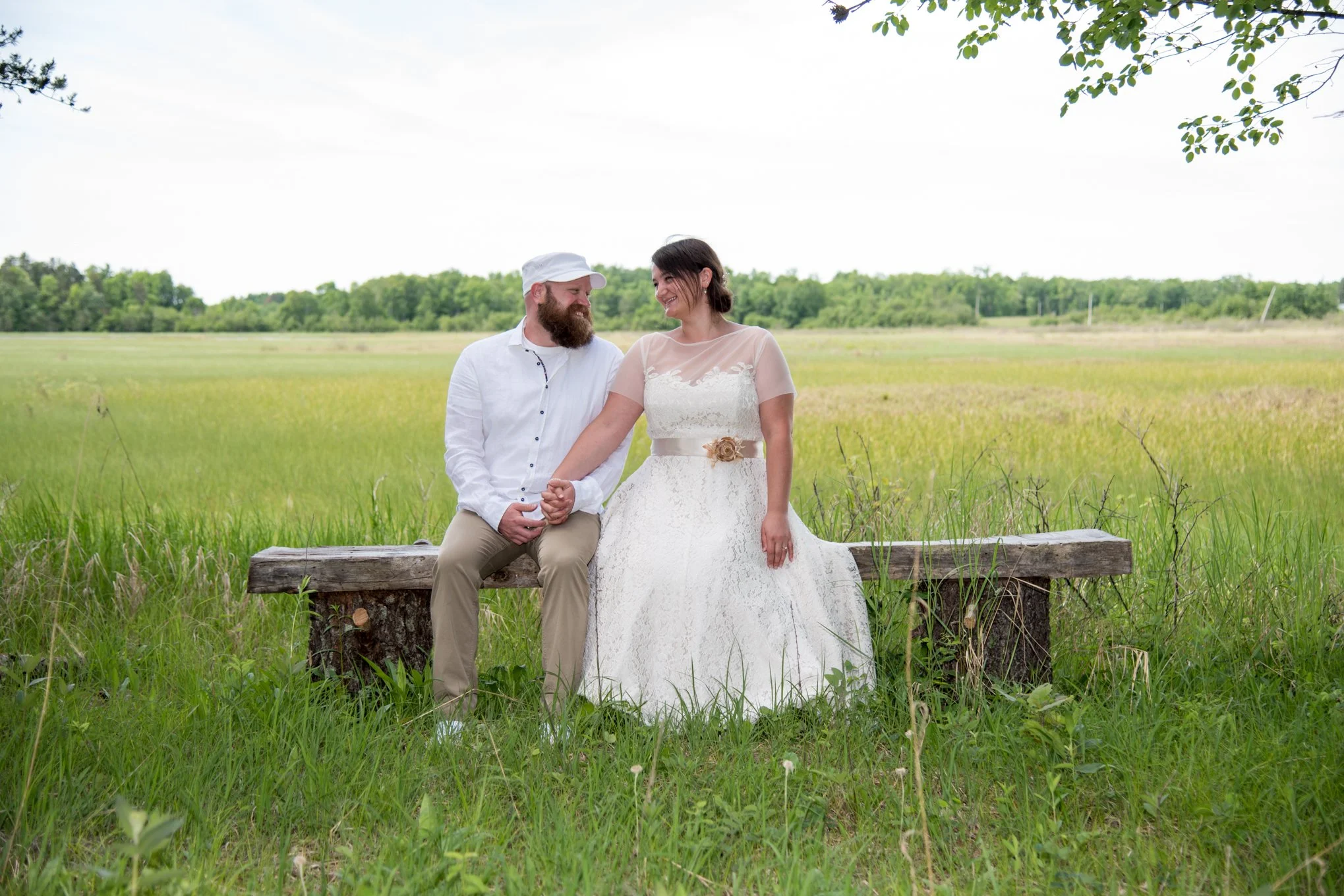 A couple dressed in wedding attire sitting on a wooden bench outdoors in a grassy field, holding hands and smiling at each other.