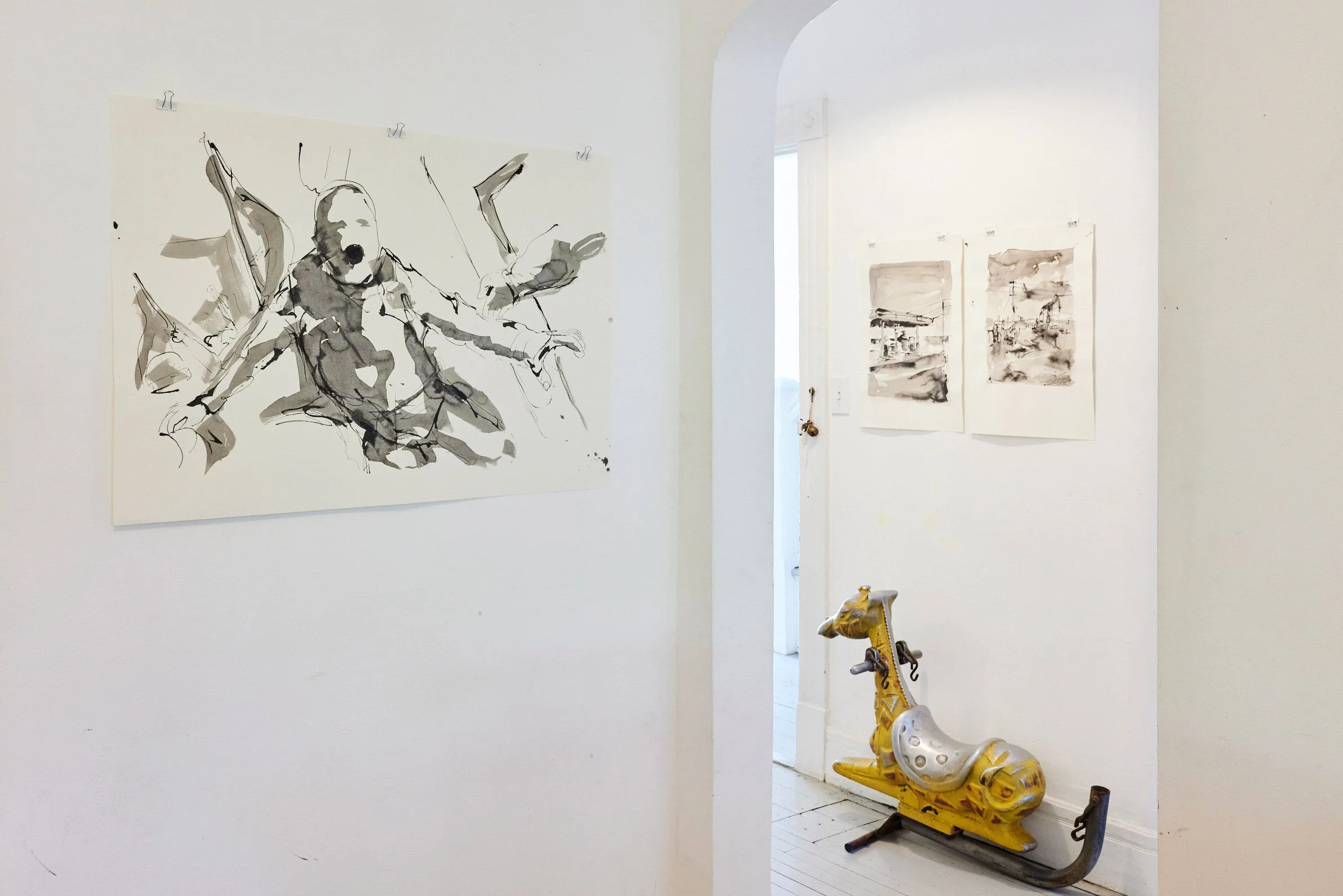 Art gallery with black and white watercolor paintings on white walls and a yellow vintage rocking horse on the floor.