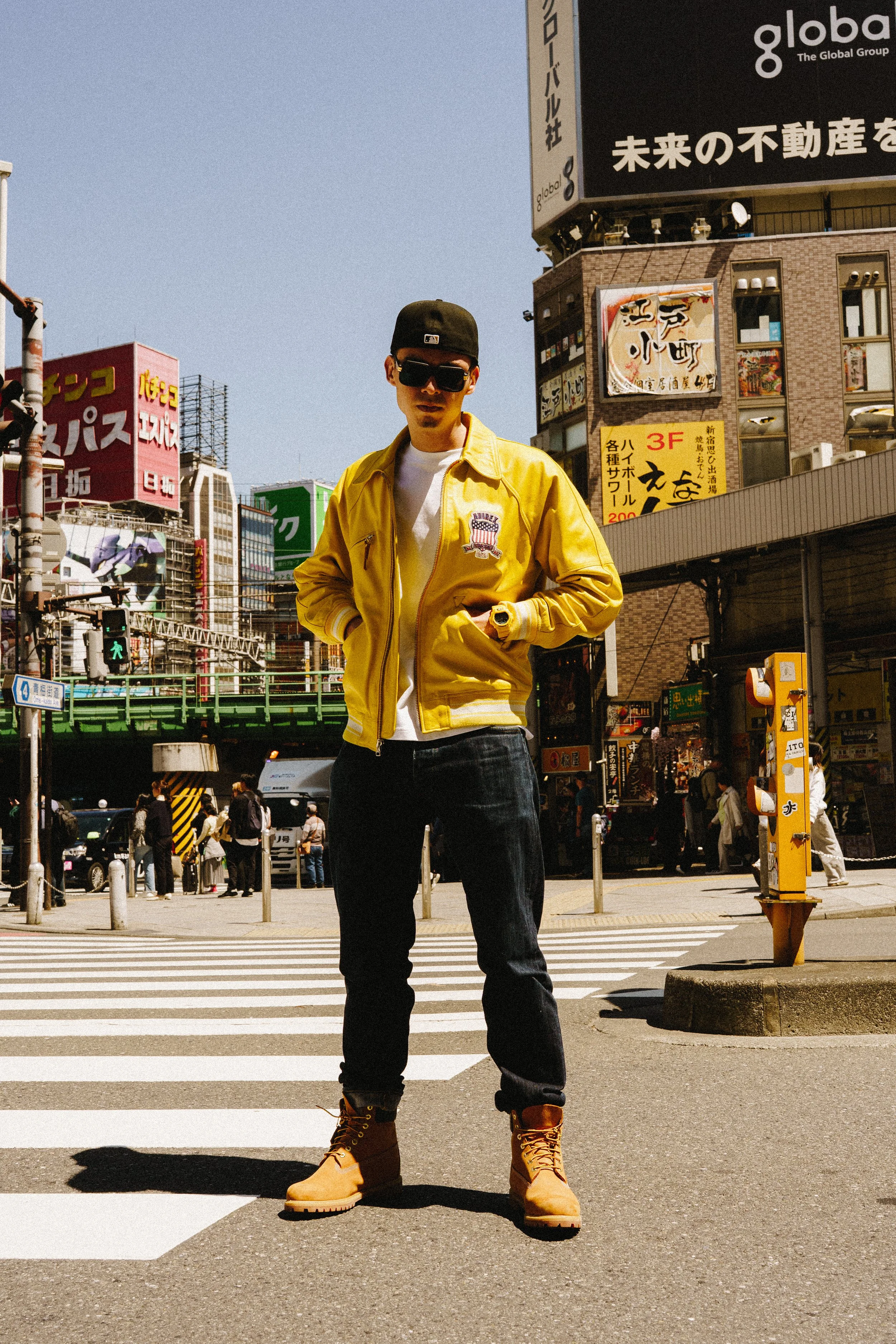 A man standing on a crosswalk in an urban area with lots of colorful signs and buildings, wearing a yellow jacket, dark jeans, tan boots, sunglasses, and a black cap in Tokyo.