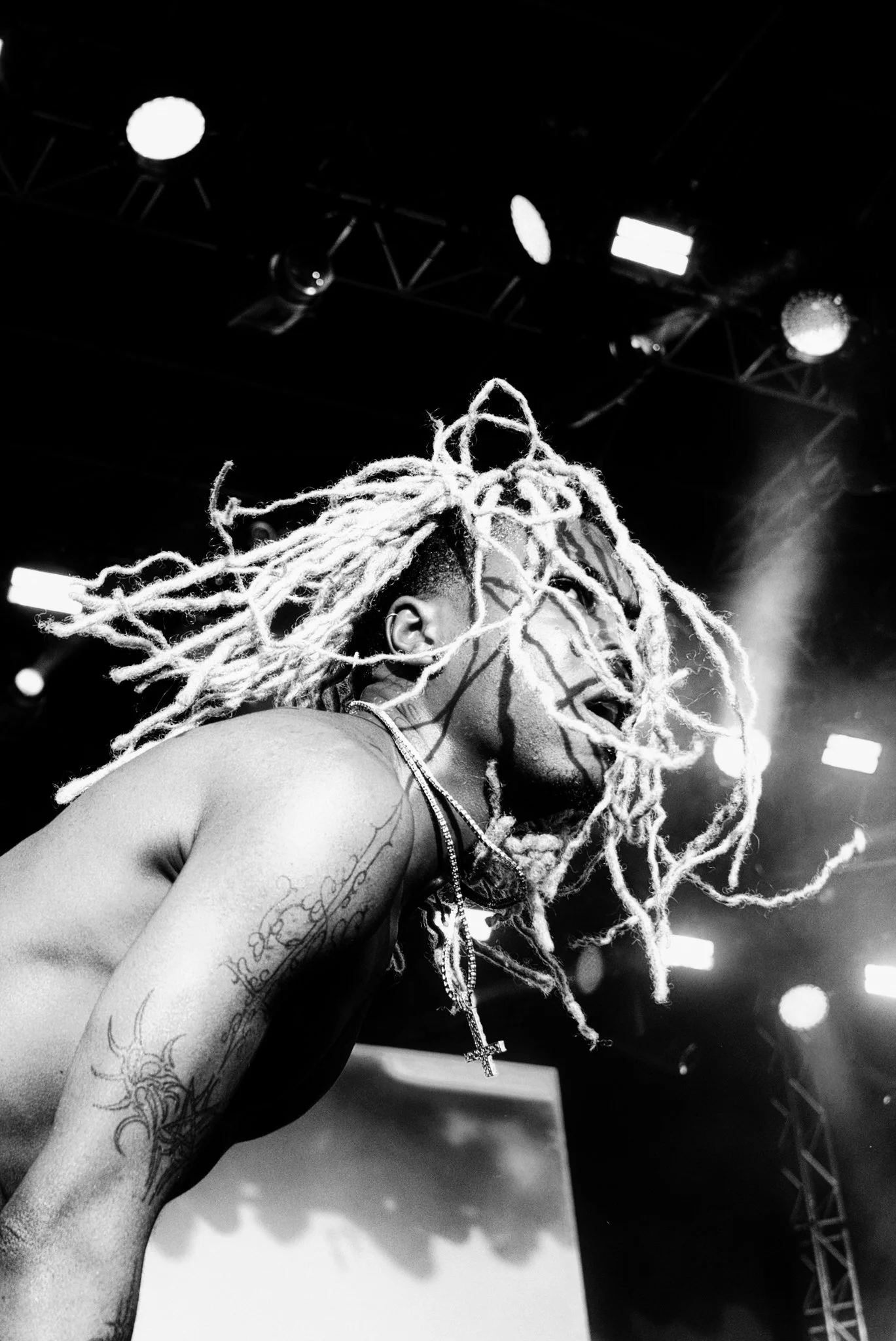A black and white photo of rapper Destroy Lonely with dreadlocks on stage, seen from a low angle, with stage lights above.