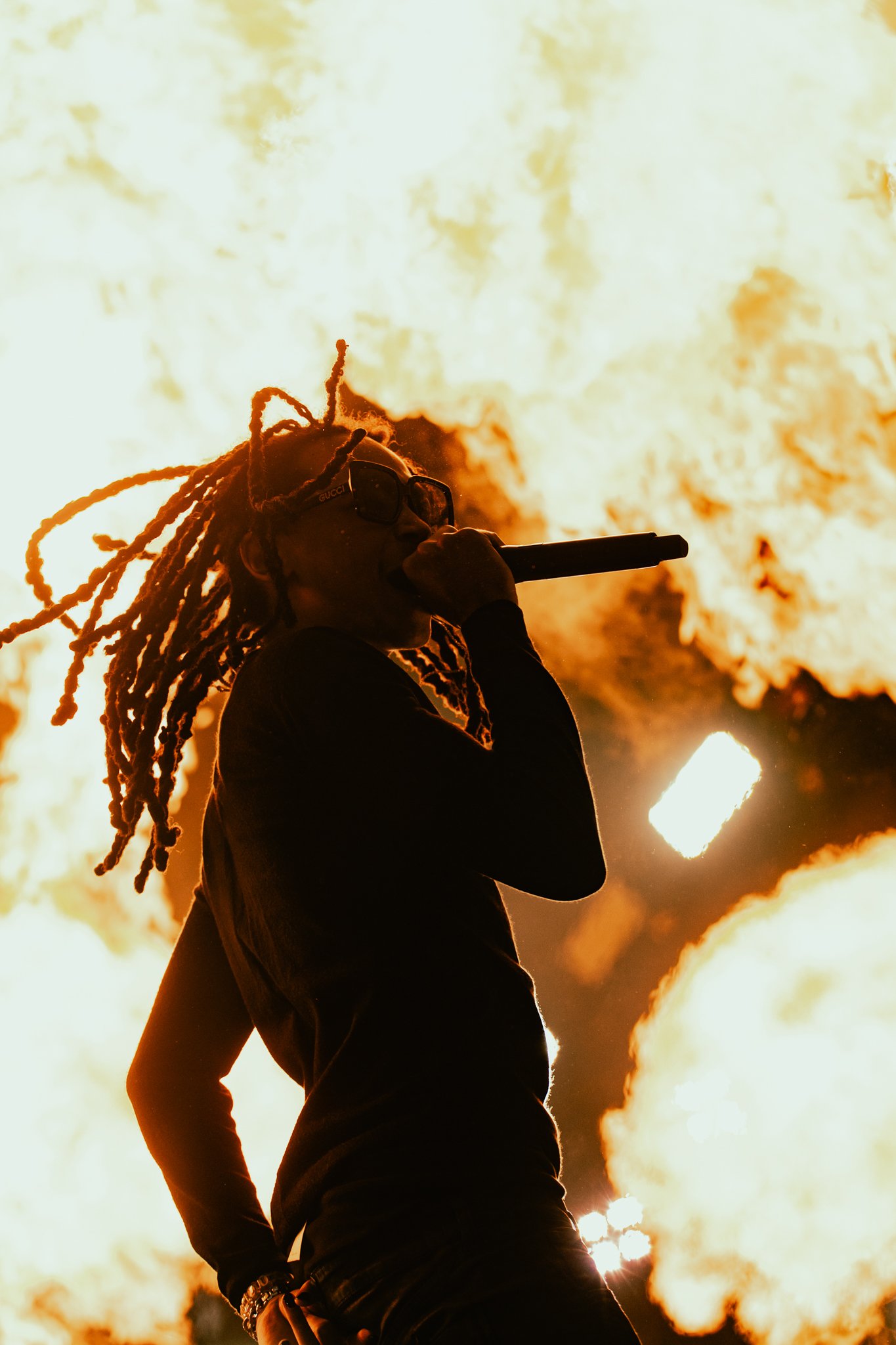 Rapper CHE wearing sunglasses holding microphone on stage with large flames and fire in the background.