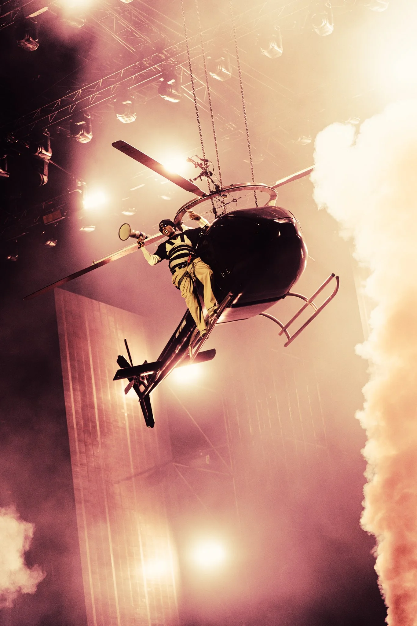 Rapper ASAP Rocky dressed in black and yellow clothing sitting on a black helicopter flying indoors with bright lights and smoke effects in the background.