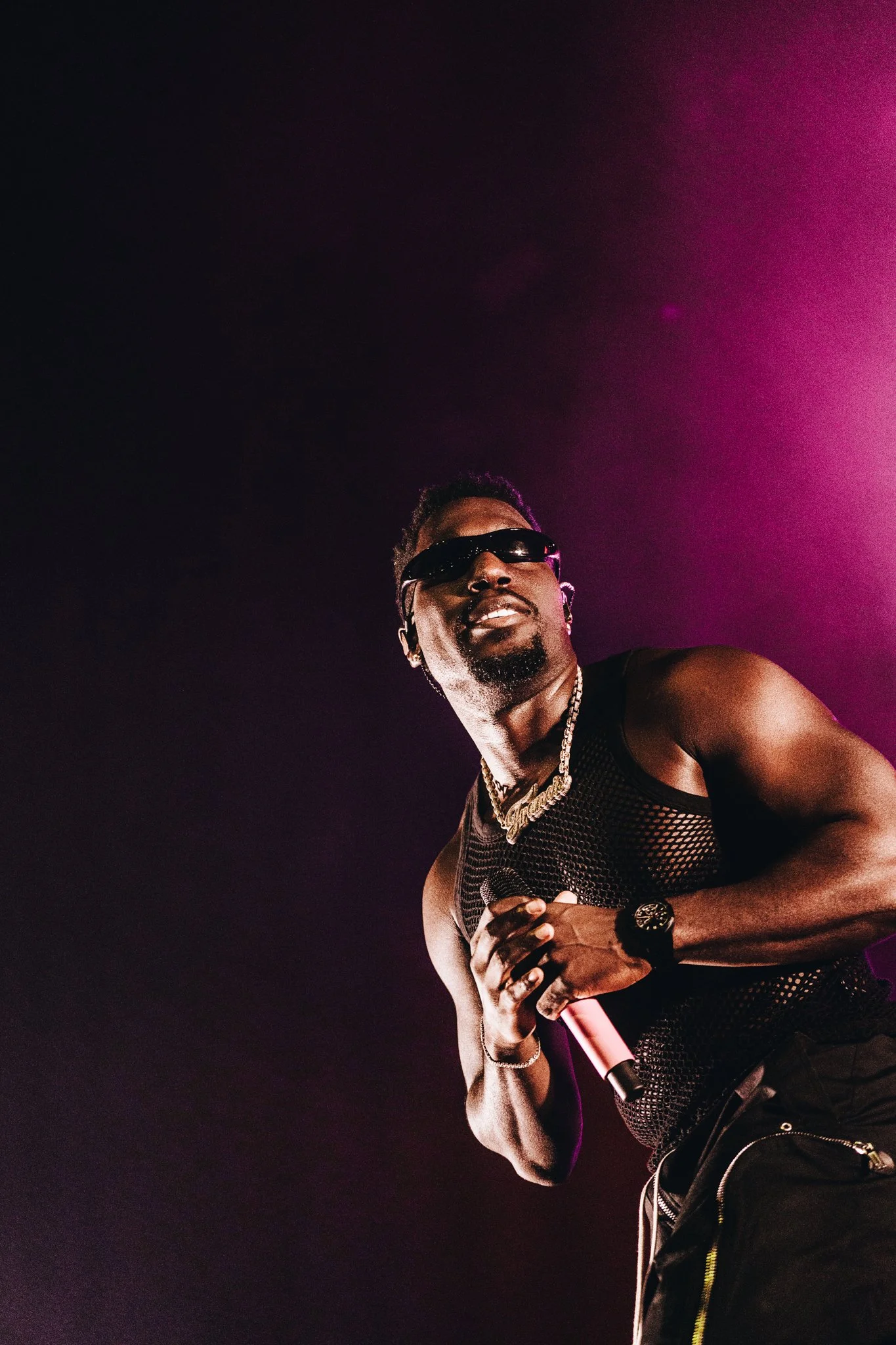 Dutch artist Frenna wearing sunglasses, a black mesh tank top, and jewelry, holding a microphone on stage with pink and purple lighting.