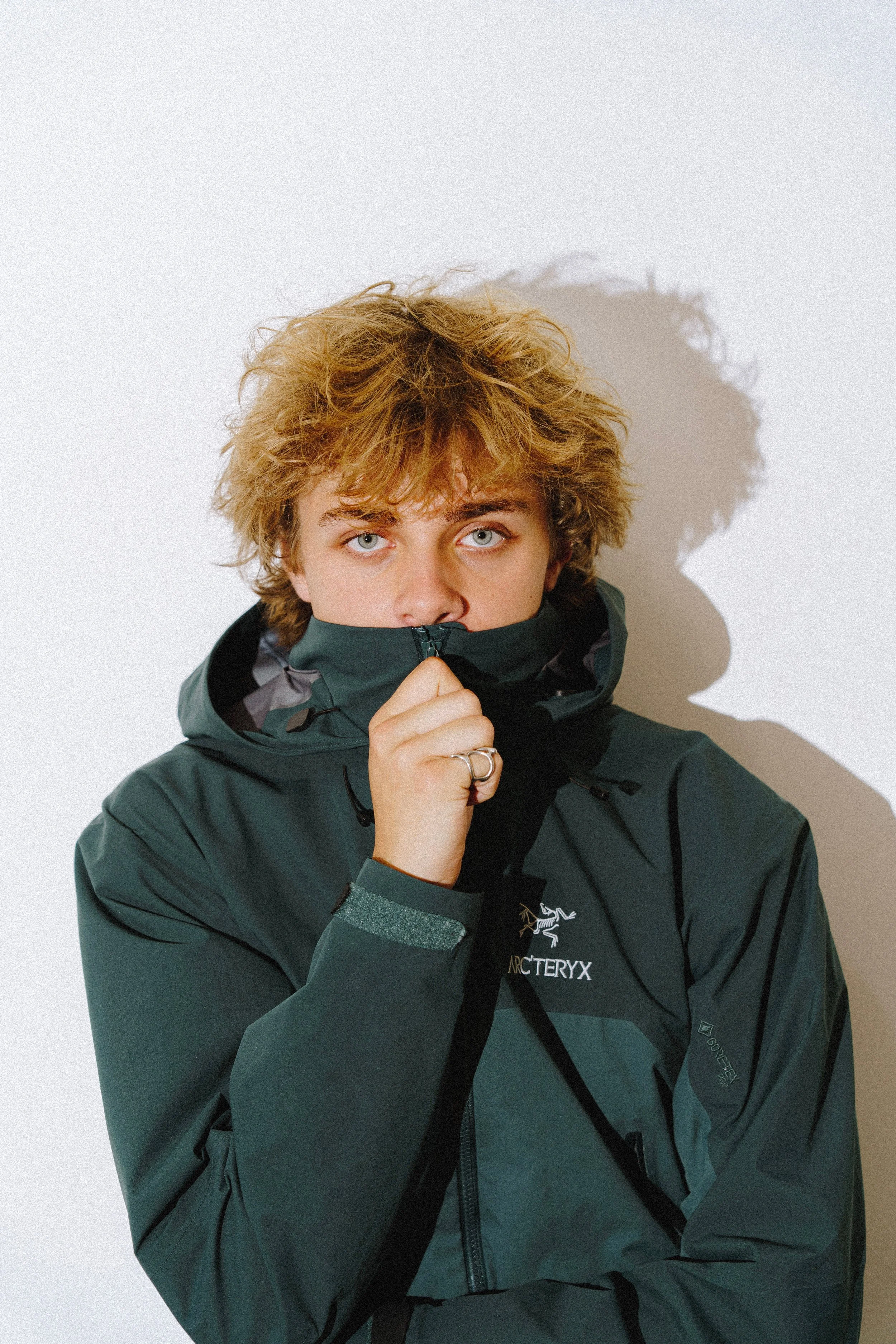 Jools Bosman with curly blond hair and blue eyes wearing a dark green Arc'teryx jacket, holding the collar up to partially cover their face, standing against a plain white background.