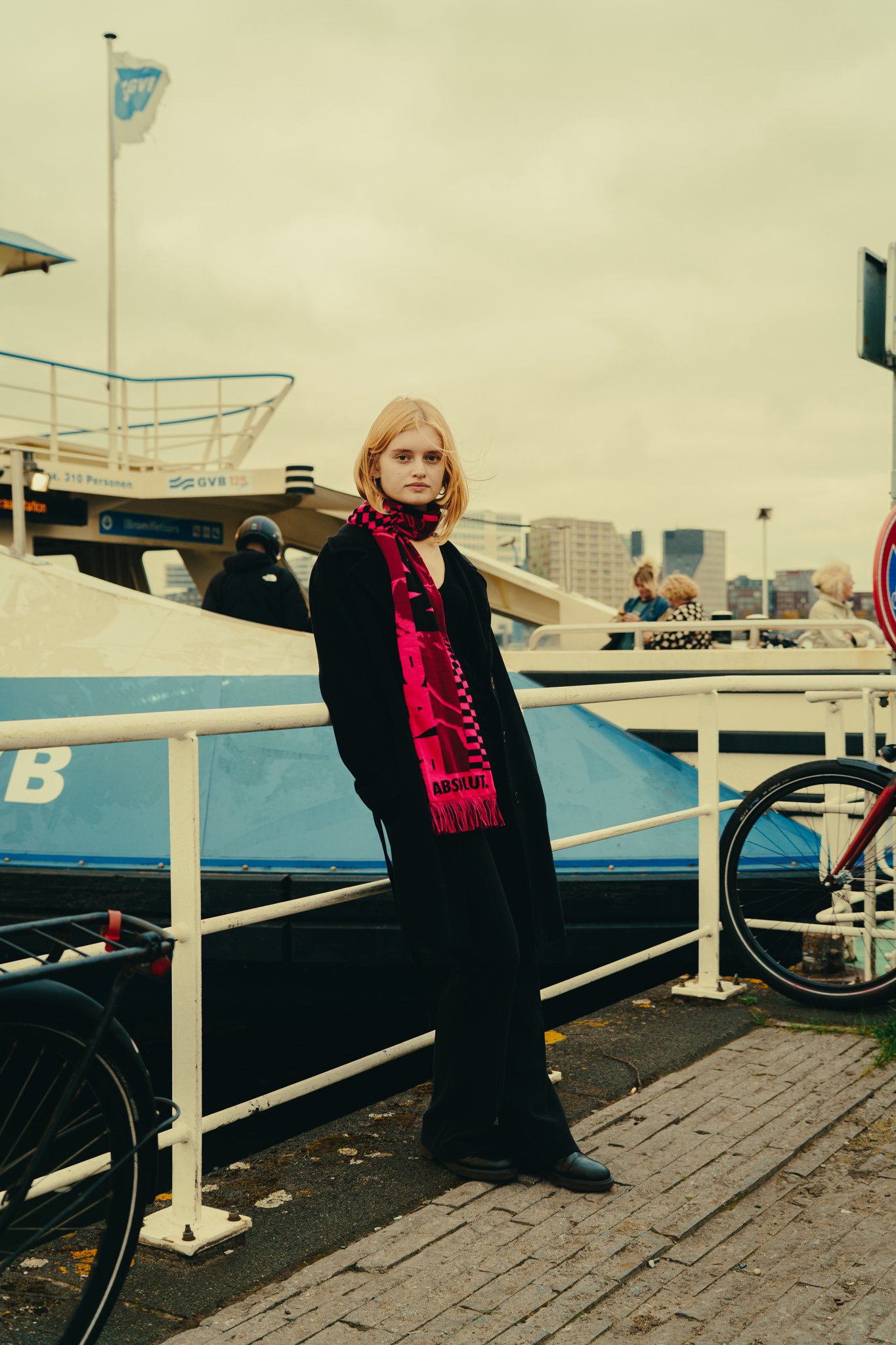 A young woman with blonde hair wearing a black coat and bright pink scarf standing on a dock near a canal with a ship in the background and other people walking by.