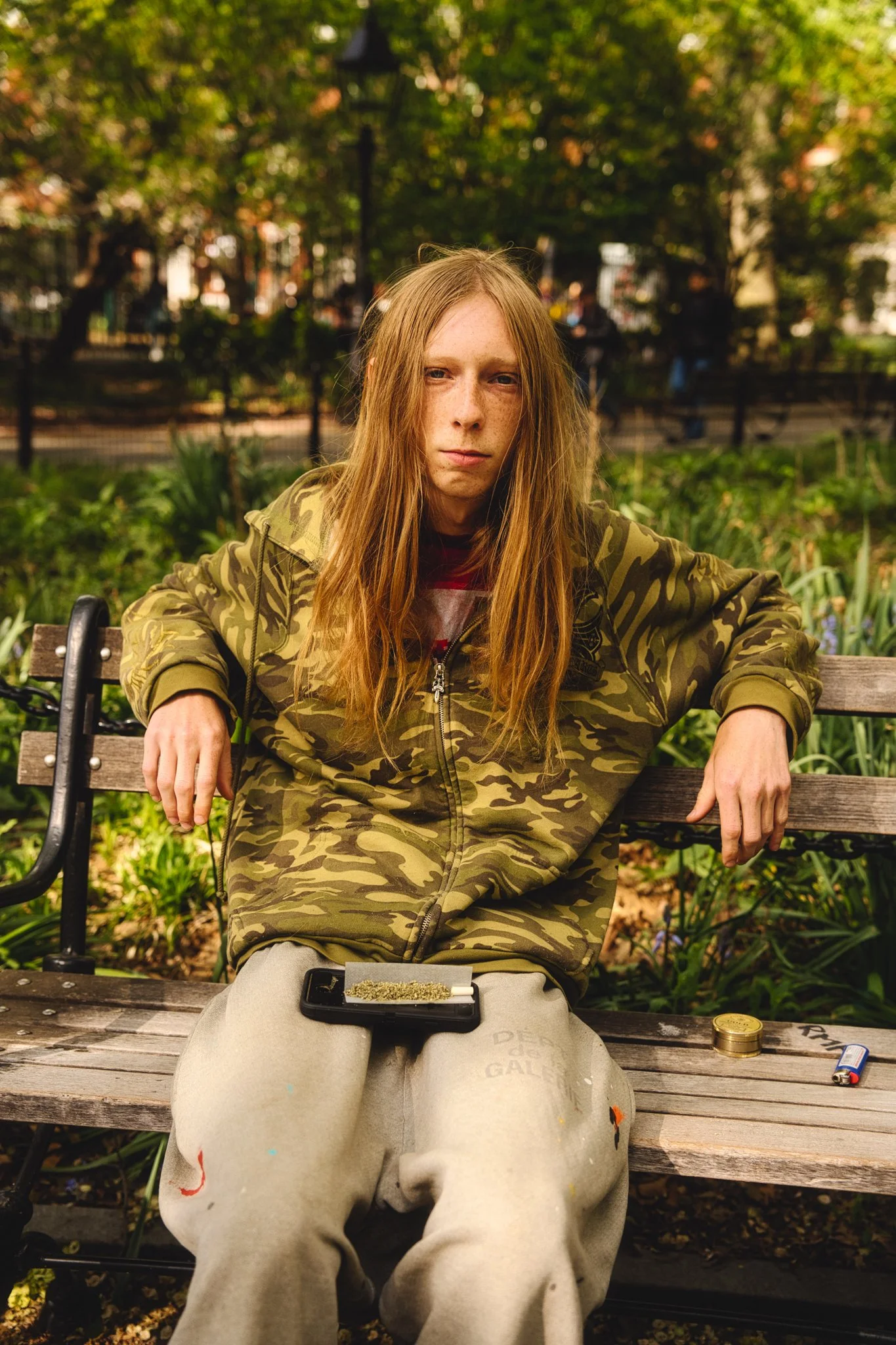 A young woman with long red hair and a serious expression sits on a park bench, wearing a camouflage jacket and beige pants. There is a marijuana joint and a phone with dried cannabis on it resting on her lap, with coins and a lighter on the bench be