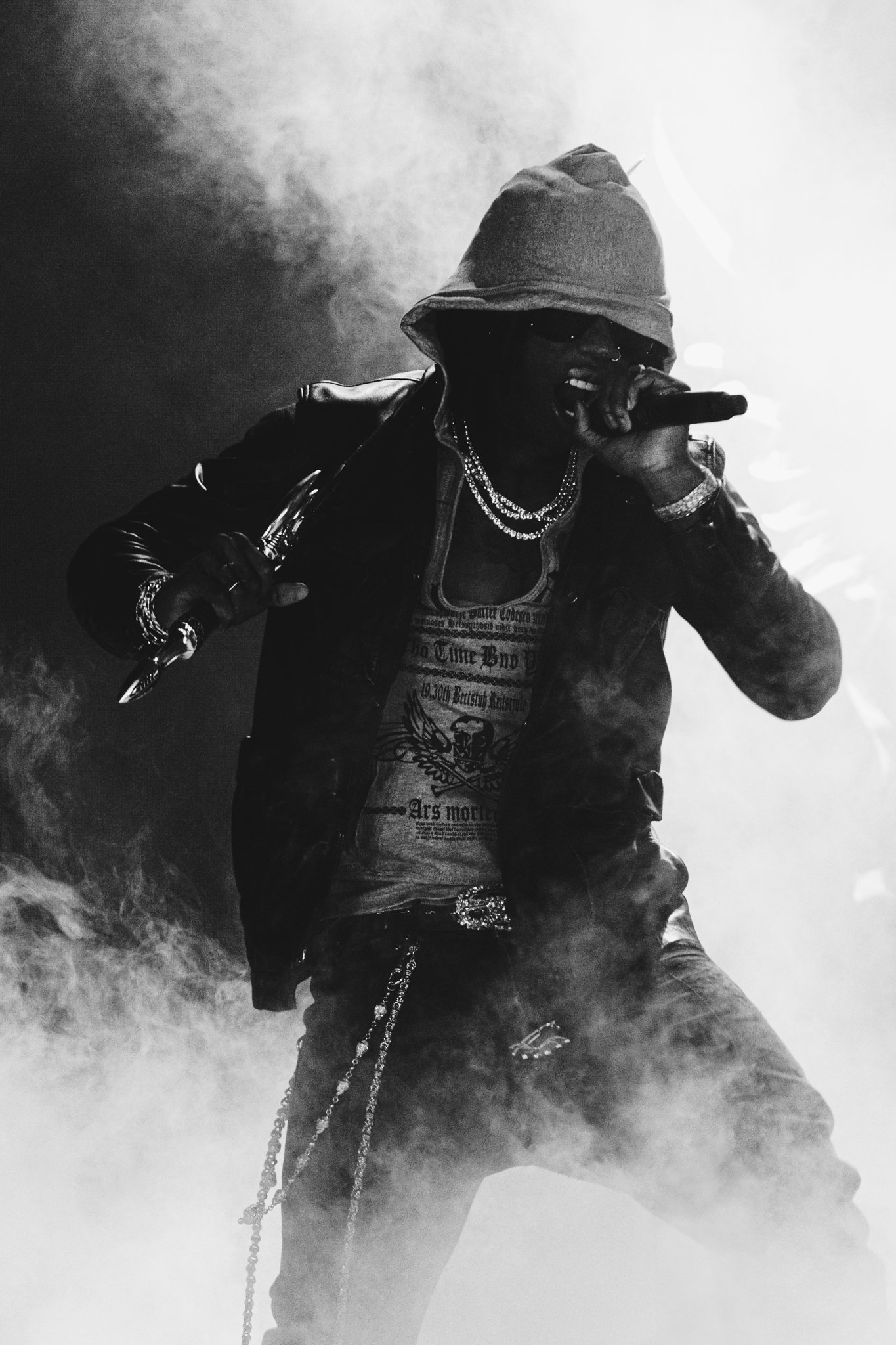 Artists Rema wearing a hoodie, sunglasses, and multiple chains, singing into a microphone on stage with smoke and fog effects.