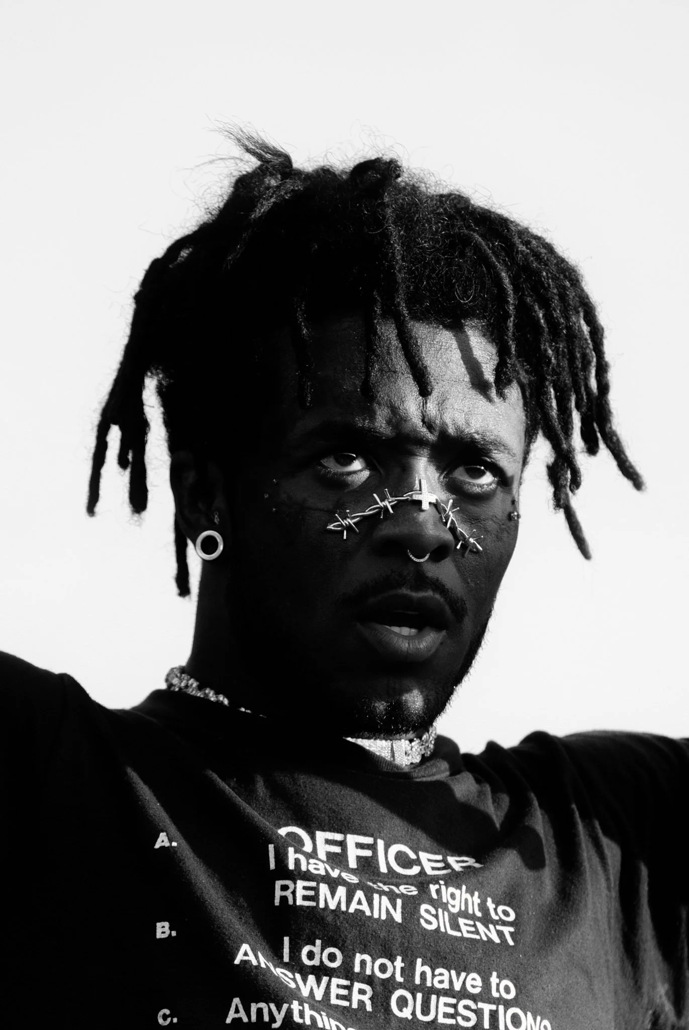 Rapper Lil Uzi Vert, wearing multiple facial piercings, jewelry, and a T-shirt with text. He has an intense expression.