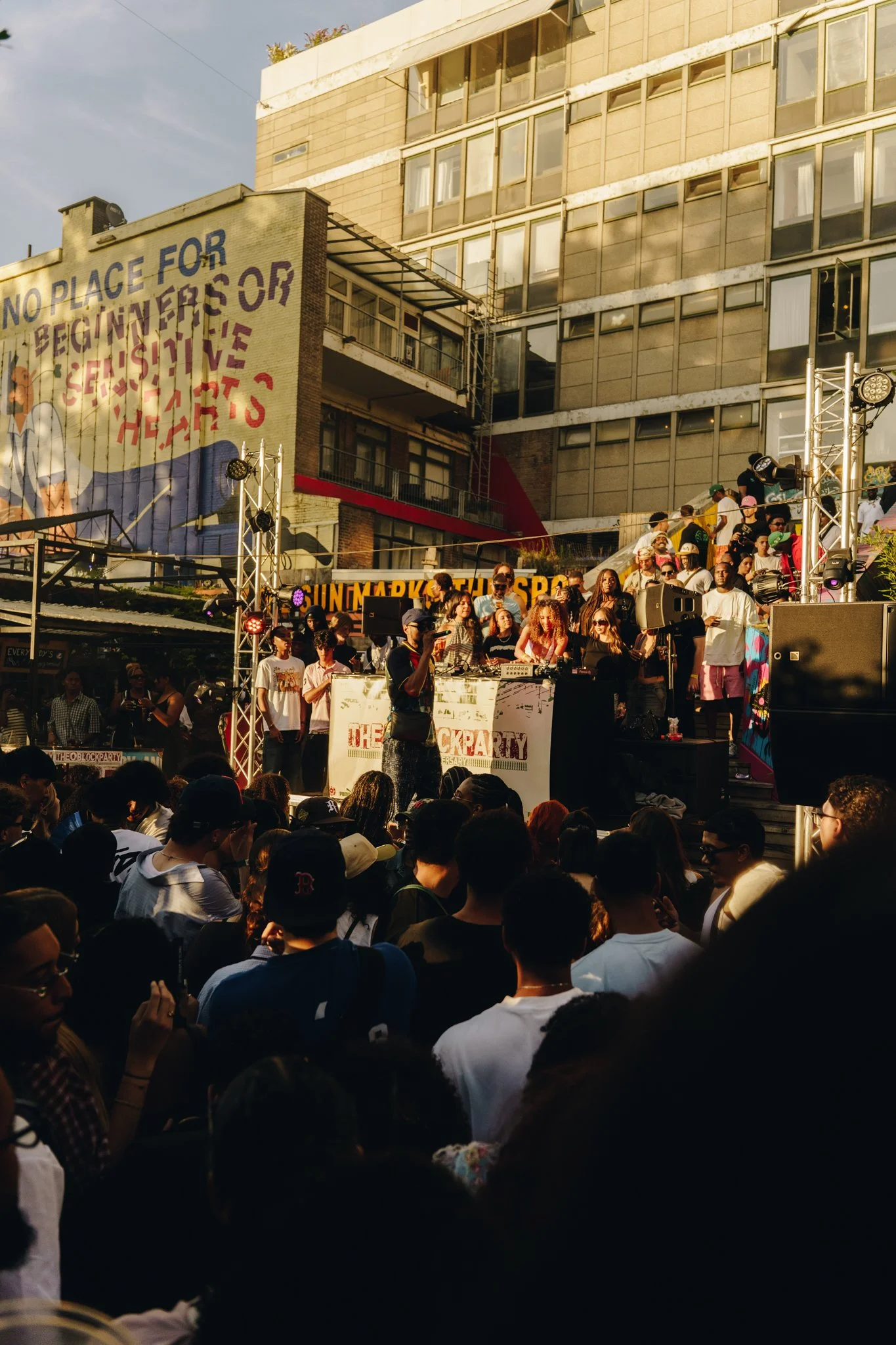 Crowd gathered at an outdoor event with a stage, where a DJ is performing. The background shows a multi-story building with a large mural and some signage.