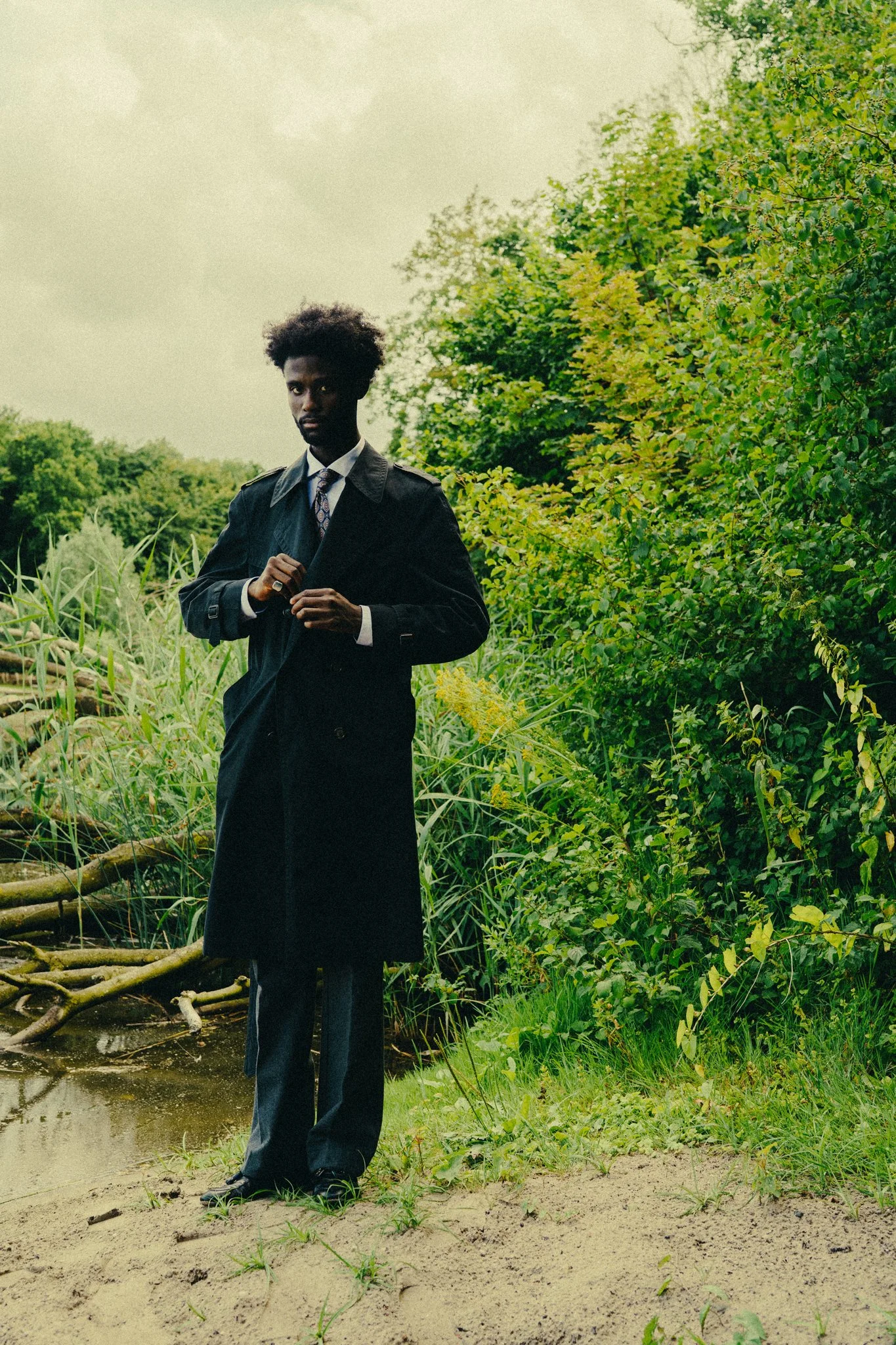 A man in a black trench coat, suit, and tie standing on a sandy patch near a body of water, surrounded by green bushes and trees, with an overcast sky in the background.