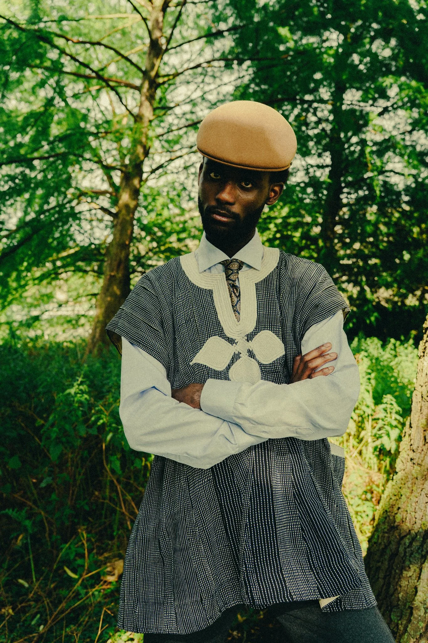 A man wearing a beige beret, a white shirt, and a patterned vest stands outdoors with his arms crossed, in front of a green, wooded background.
