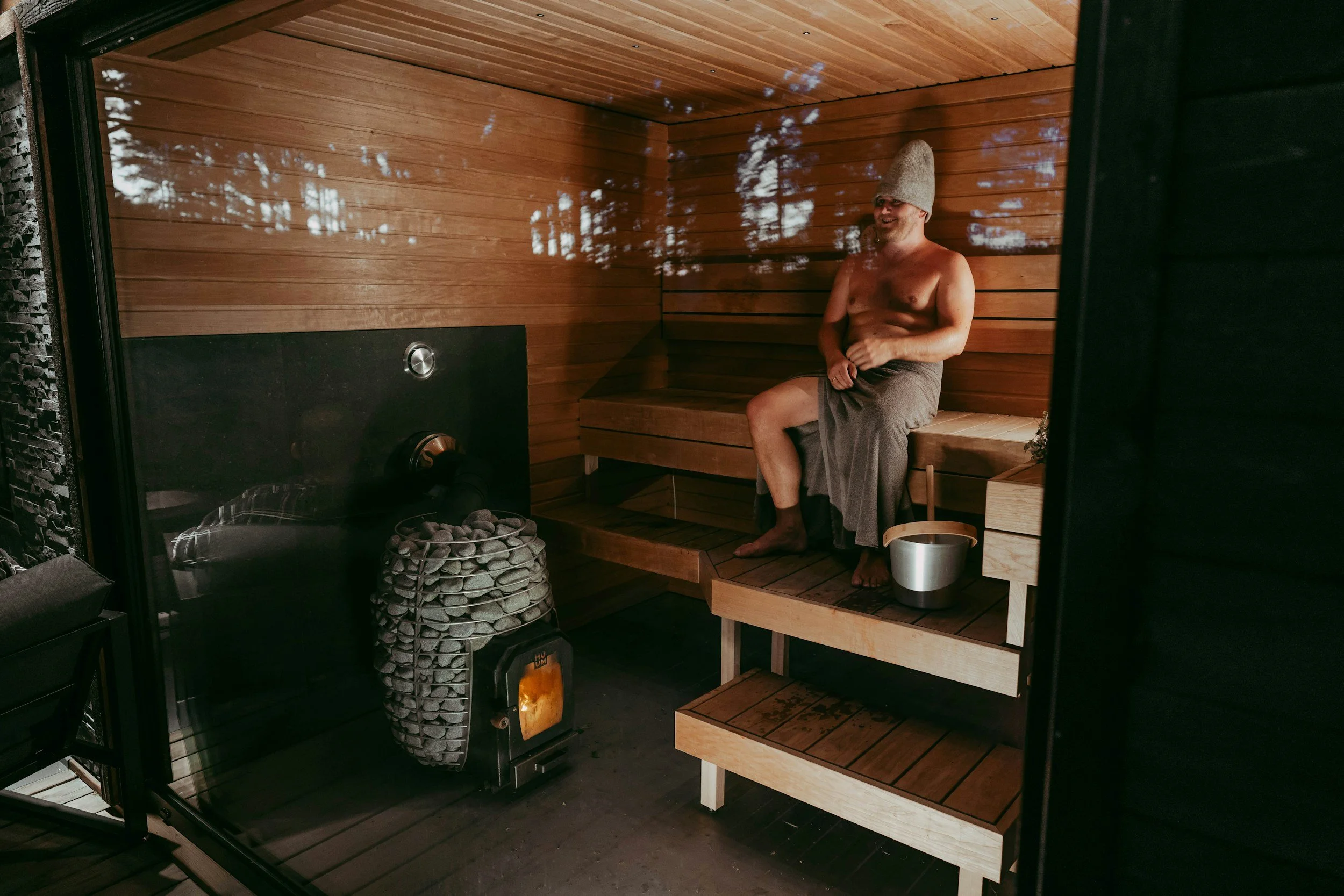 A man sitting shirtless in a wooden sauna, wearing a towel and a gray hat, smiling. There is a bucket with a ladle beside him and a small heater with stones and a stove in the corner.