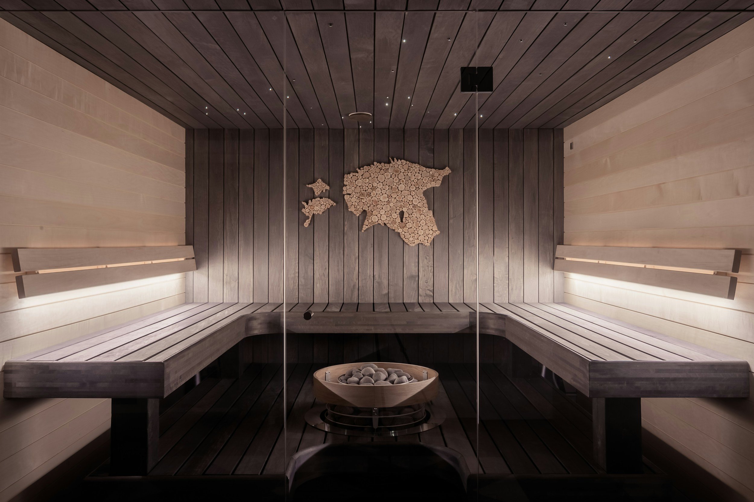 Interior of a sauna with wooden benches and walls, and a decorative wooden map of Greece on the back wall, with a bench and a stone heater in the foreground.