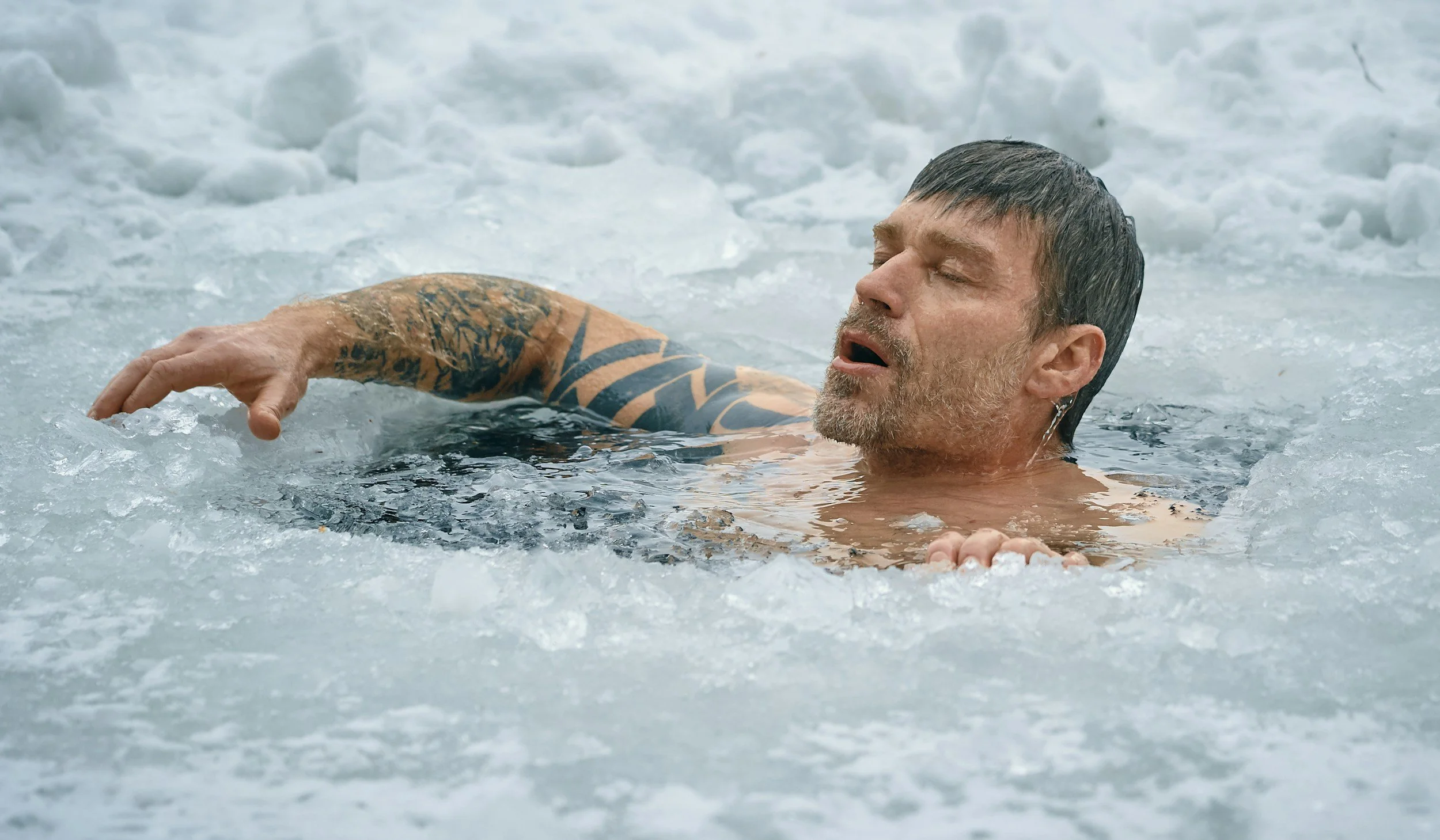 A man with tattoos on his arm is swimming in icy water with ice and snow around him, appearing to be exhausted or in distress.
