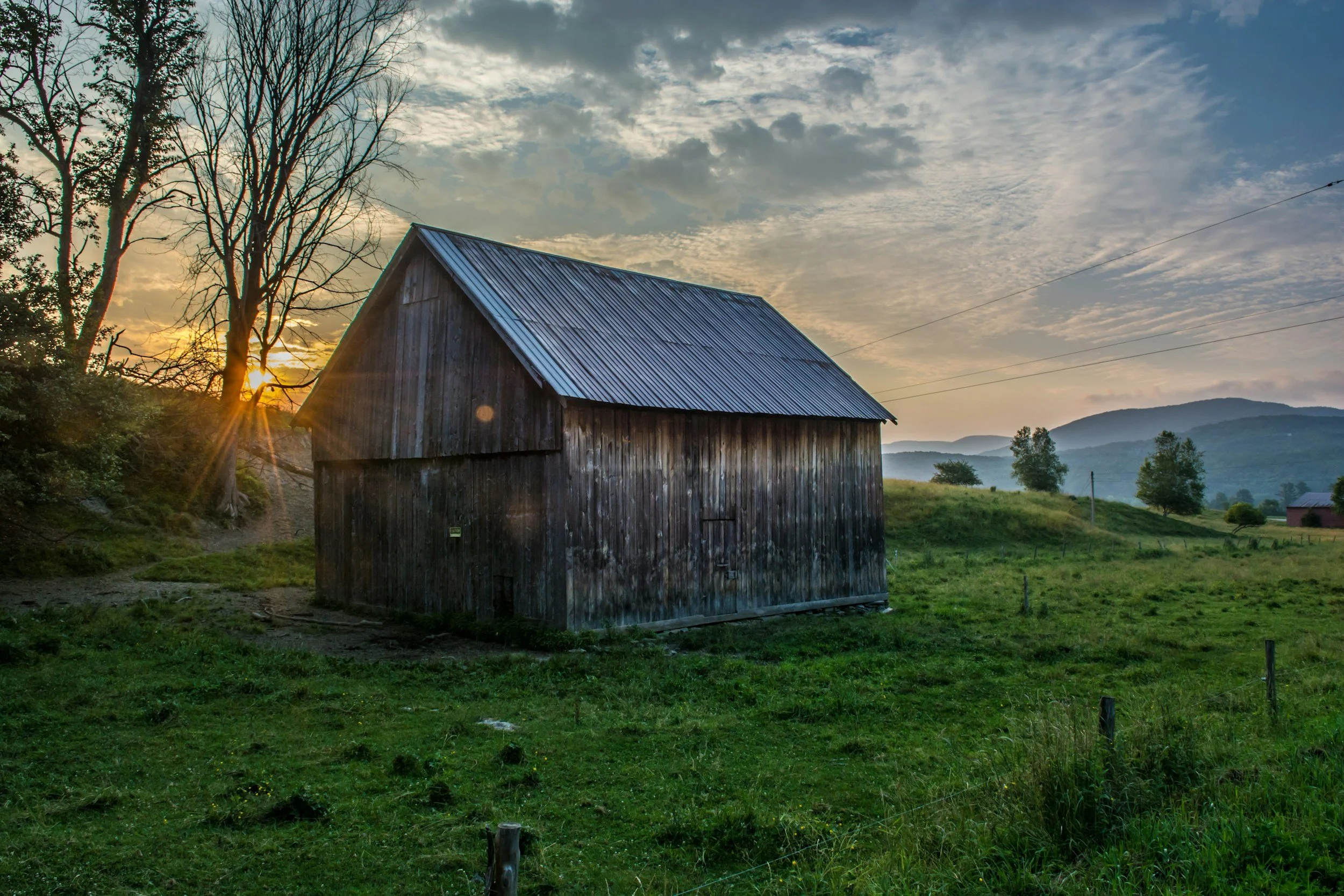 A rustic wooden barn in a grassy field during sunset, with a tree and mountain in the background.