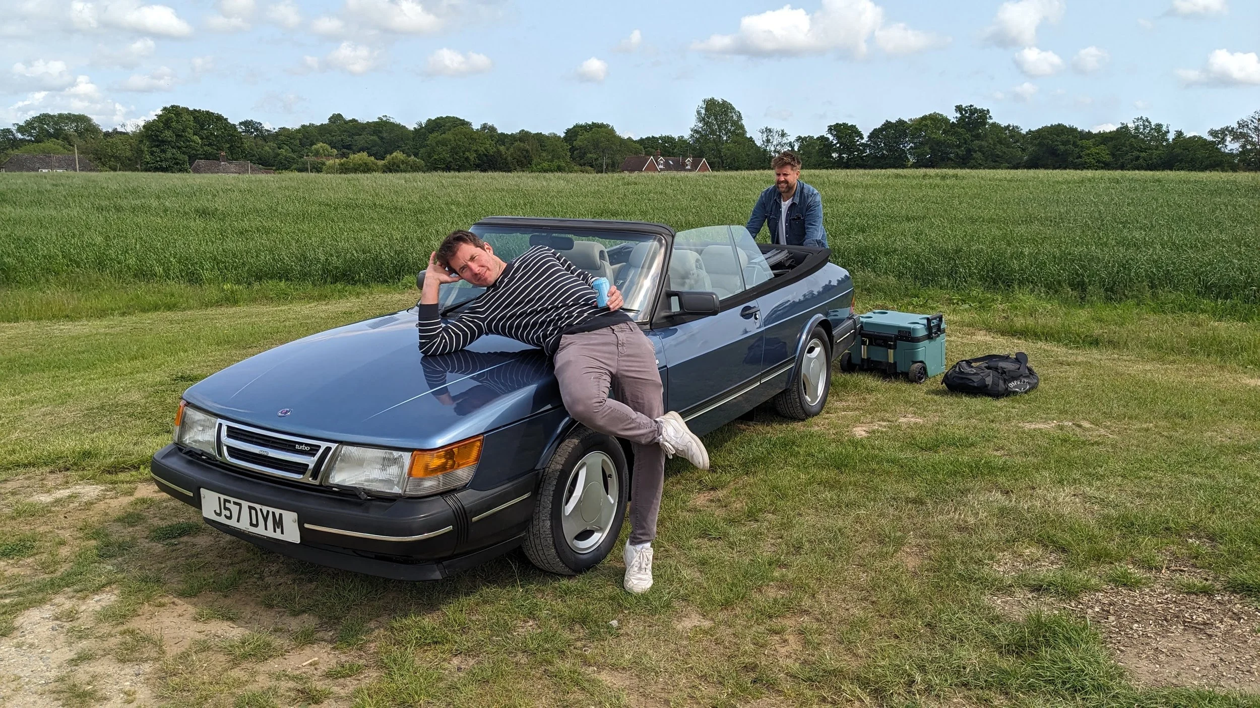Two men with a blue convertible car on a grassy field, one lying on the car's hood and another standing nearby, with outdoor equipment on the ground and trees in the background.