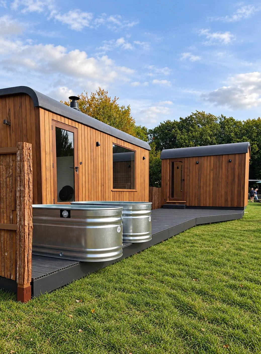 Two modern wooden tiny houses on a black deck, with smaller one in front, large sky with clouds, lush green trees, and grassy lawn in the background.