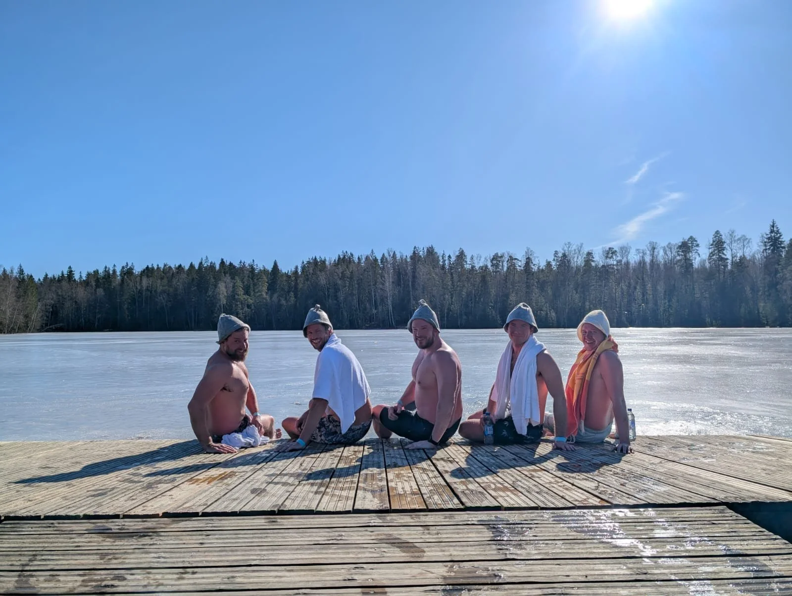 Five men sitting on a wooden dock by a frozen lake, wearing winter hats and towels, enjoying a winter swim on a sunny day.