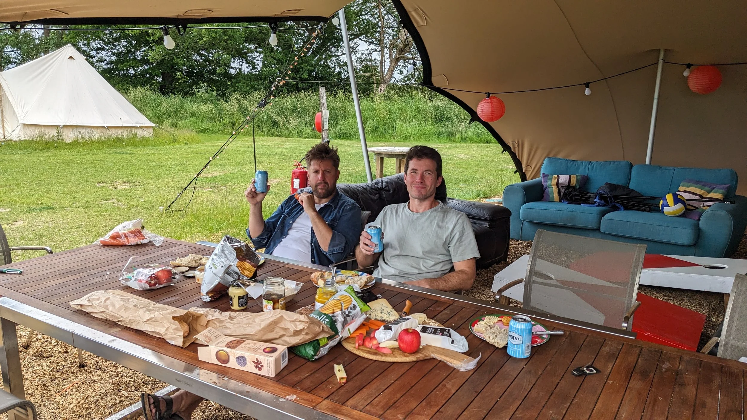 Two men sitting under a large canopy tent at a picnic table with food and drinks, in an outdoor grassy area with a white tent and trees in the background, during daytime.