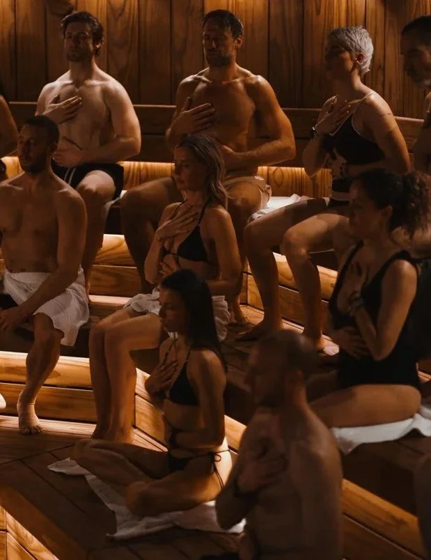 People in a dimly lit room sitting and kneeling on wooden benches, performing a yoga or meditation session, with their hands on their chests and eyes closed.