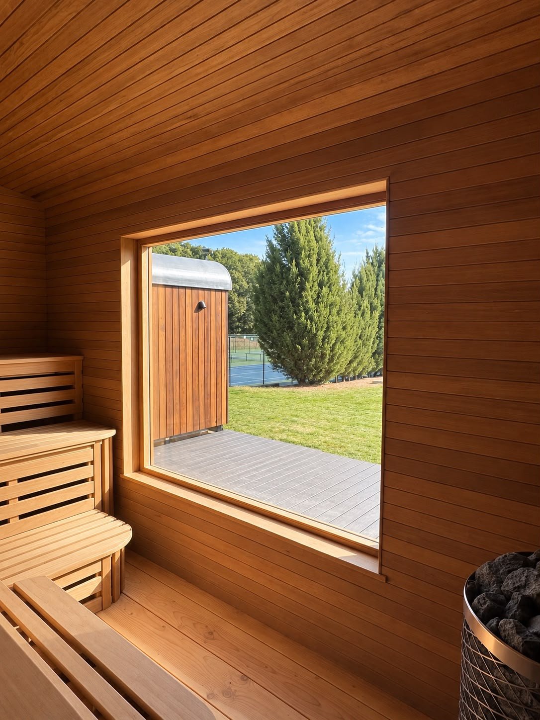 Interior of a wooden sauna with bench seating, large window opening to a grassy outdoor area with a large tree and tennis court in the background.