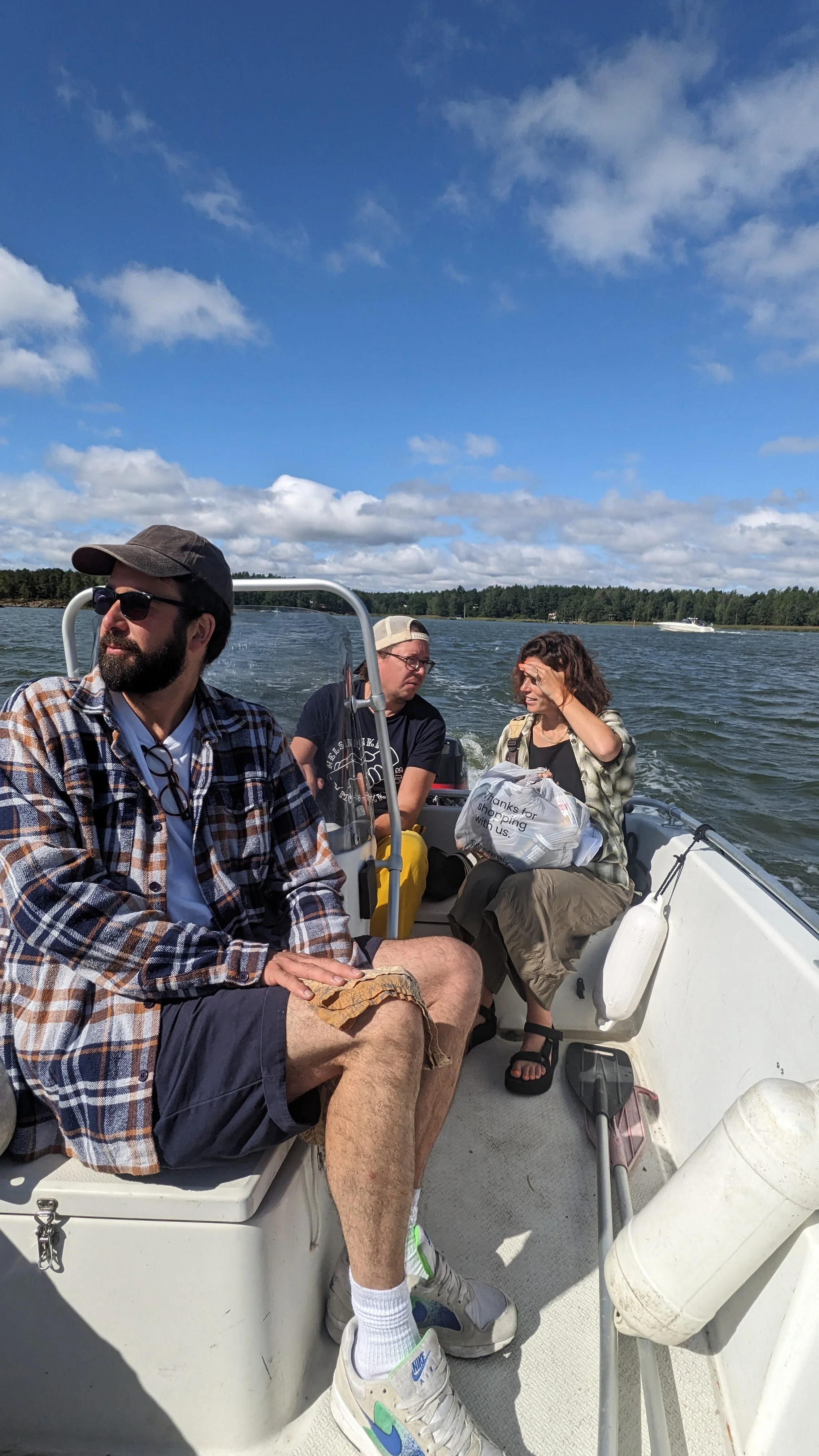 Three people sitting on a boat on a body of water with trees and partly cloudy sky in the background, one of them holding a plastic shopping bag.