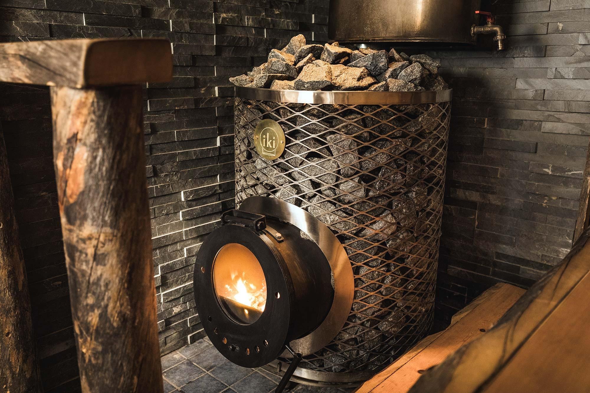 A sauna heater filled with rocks in a sauna room with dark stone walls, wood logs, and a faucet.