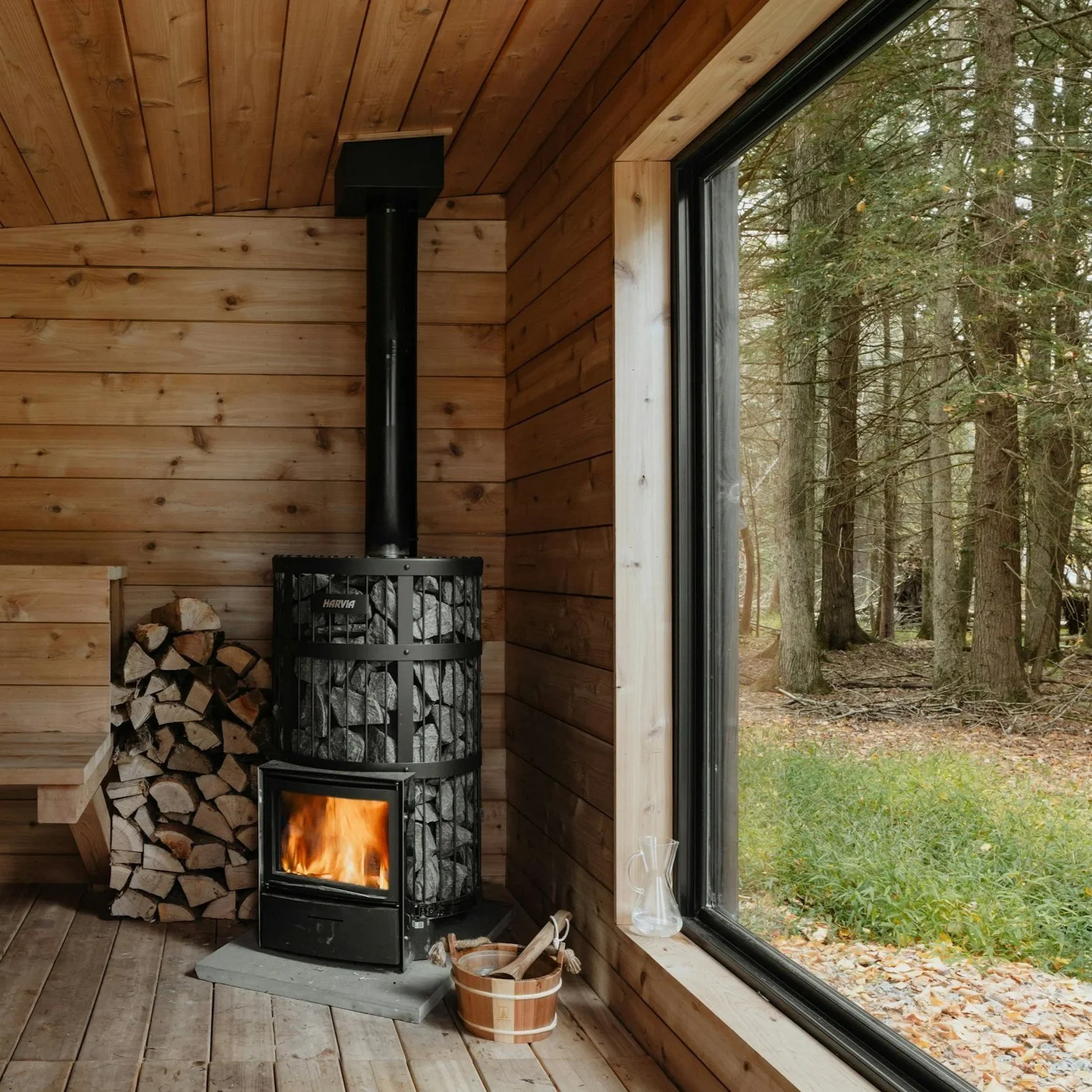 Interior of a wooden cabin with a wood stove burning, firewood stacked nearby, and large windows showing a forest outside.