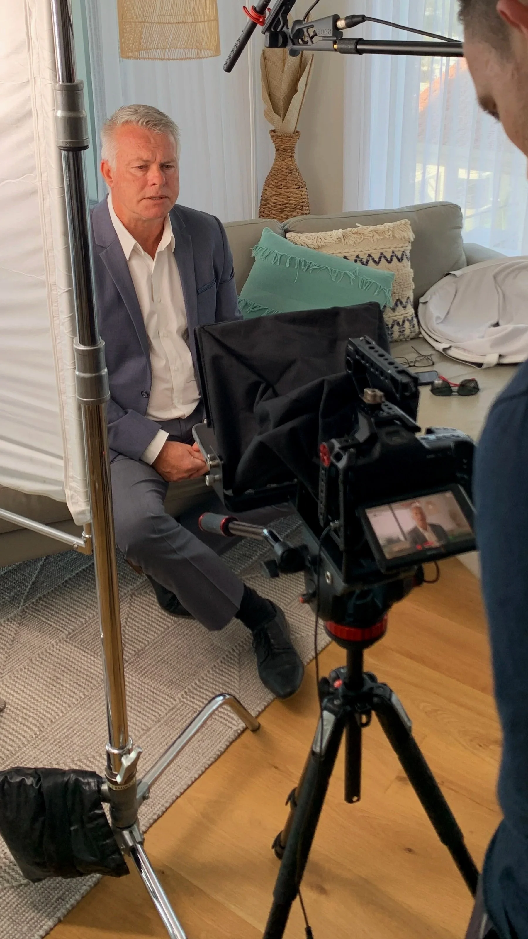 A man in a gray suit being filmed for an interview or video recording in a living room with pillows on the sofa, a lamp, and sheer curtains, with professional camera and lighting equipment.