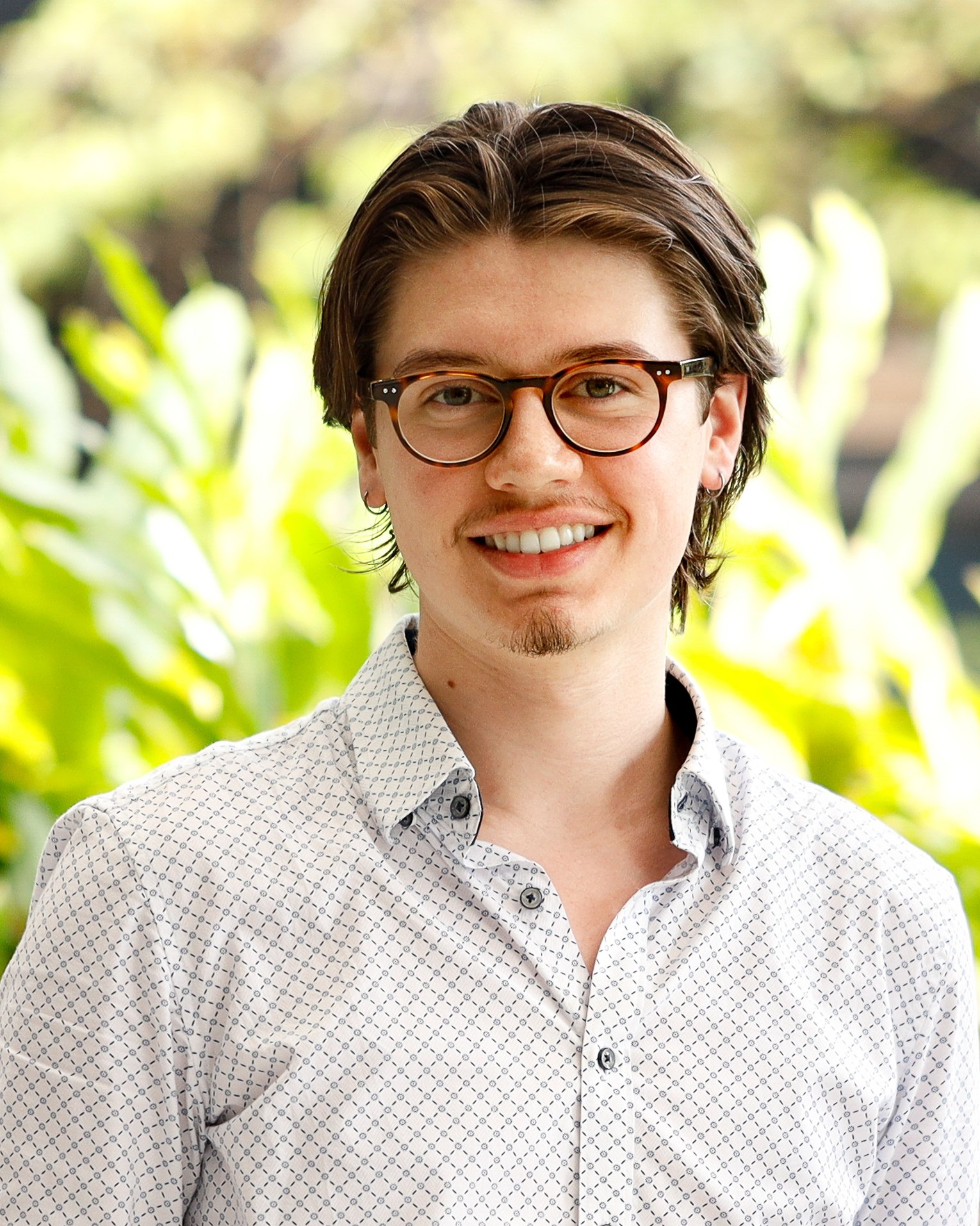 Portrait of a young man with short dark hair, glasses, and a small goatee, smiling in front of green foliage.