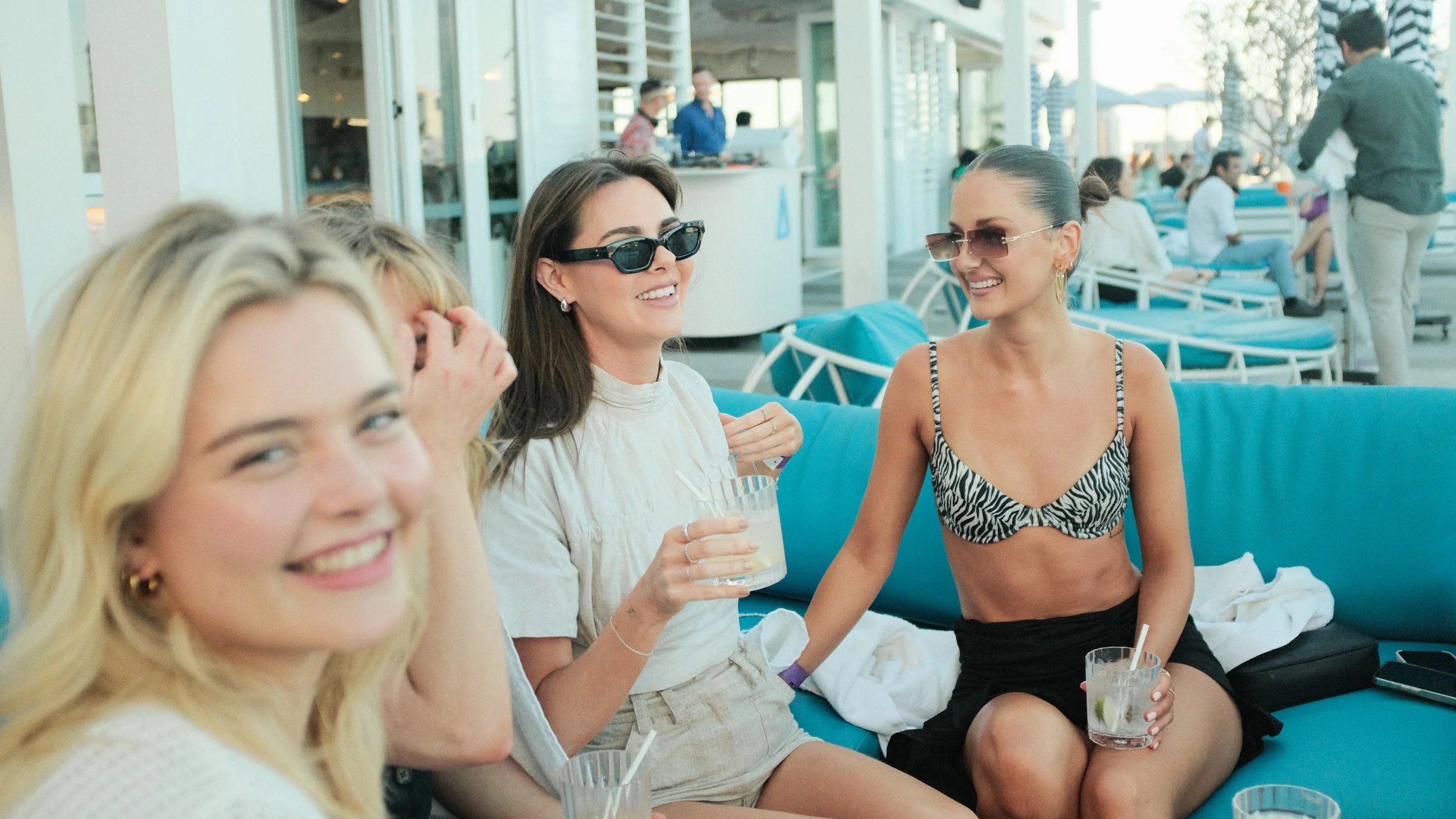 Four women sitting on a blue outdoor lounge, smiling and chatting, with drinks in hand, at a poolside or beachside venue, with other people and lounge chairs in the background.