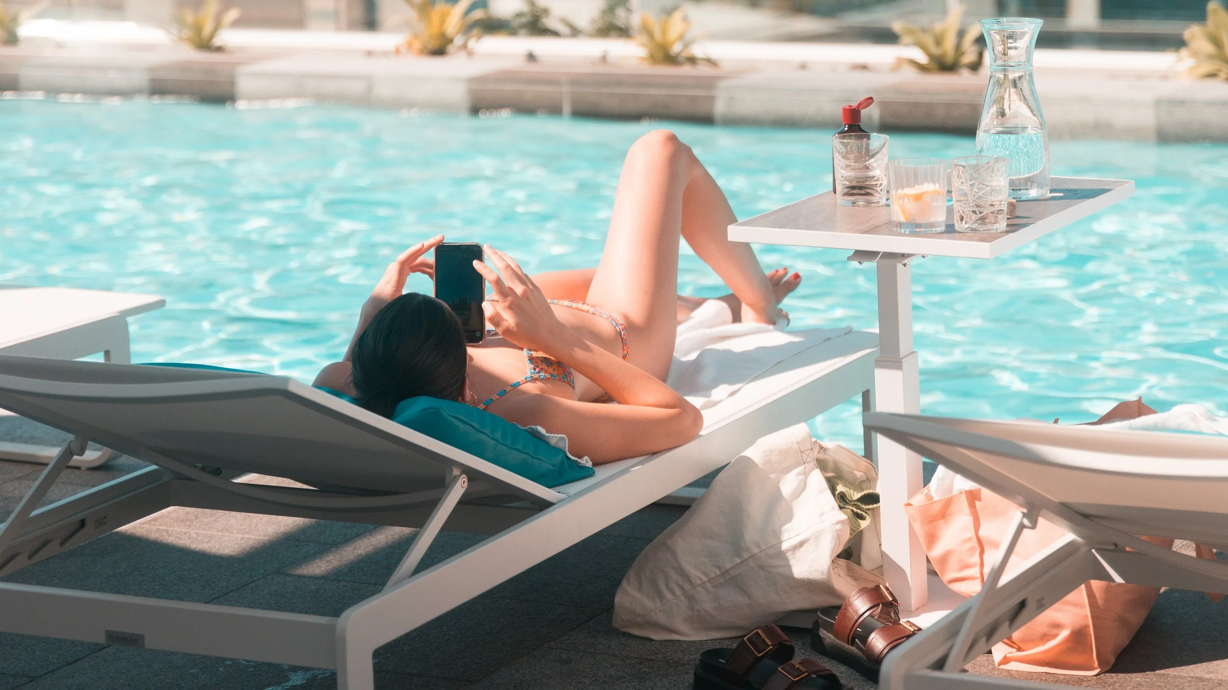 Woman relaxing on a lounge chair by a swimming pool, taking a photo with her phone. There is a small table with a water pitcher, glasses, a water bottle, and a drink. Towels, bags, and shoes are on the ground nearby.