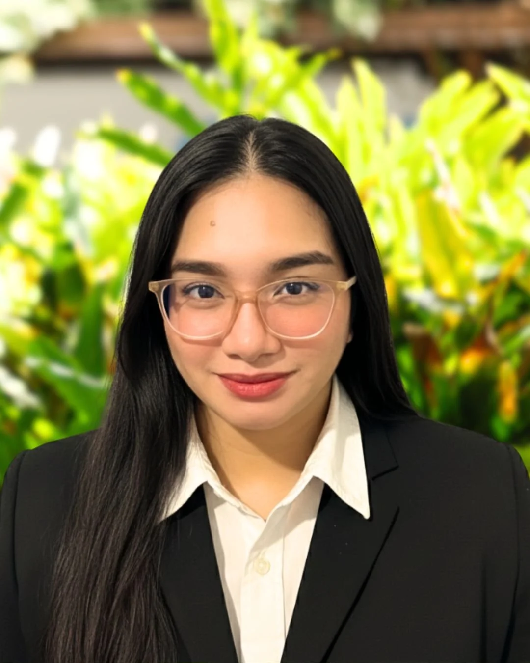 A woman with long black hair, wearing glasses, a white shirt, and a black blazer, standing in front of a green leafy background.