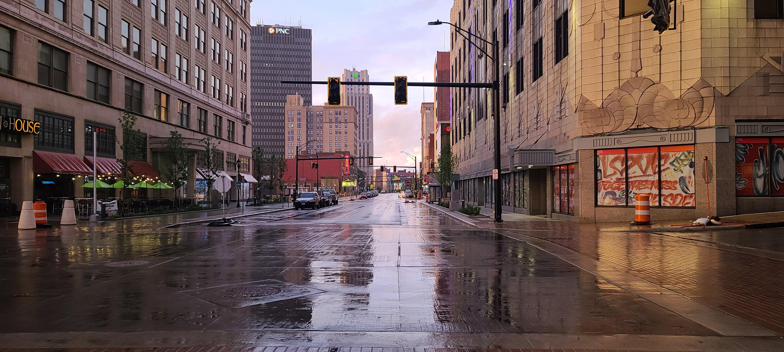 Empty city street after rain with wet pavement reflecting city lights, tall buildings, traffic lights, street signs, parked cars, and some trees along the sidewalk.