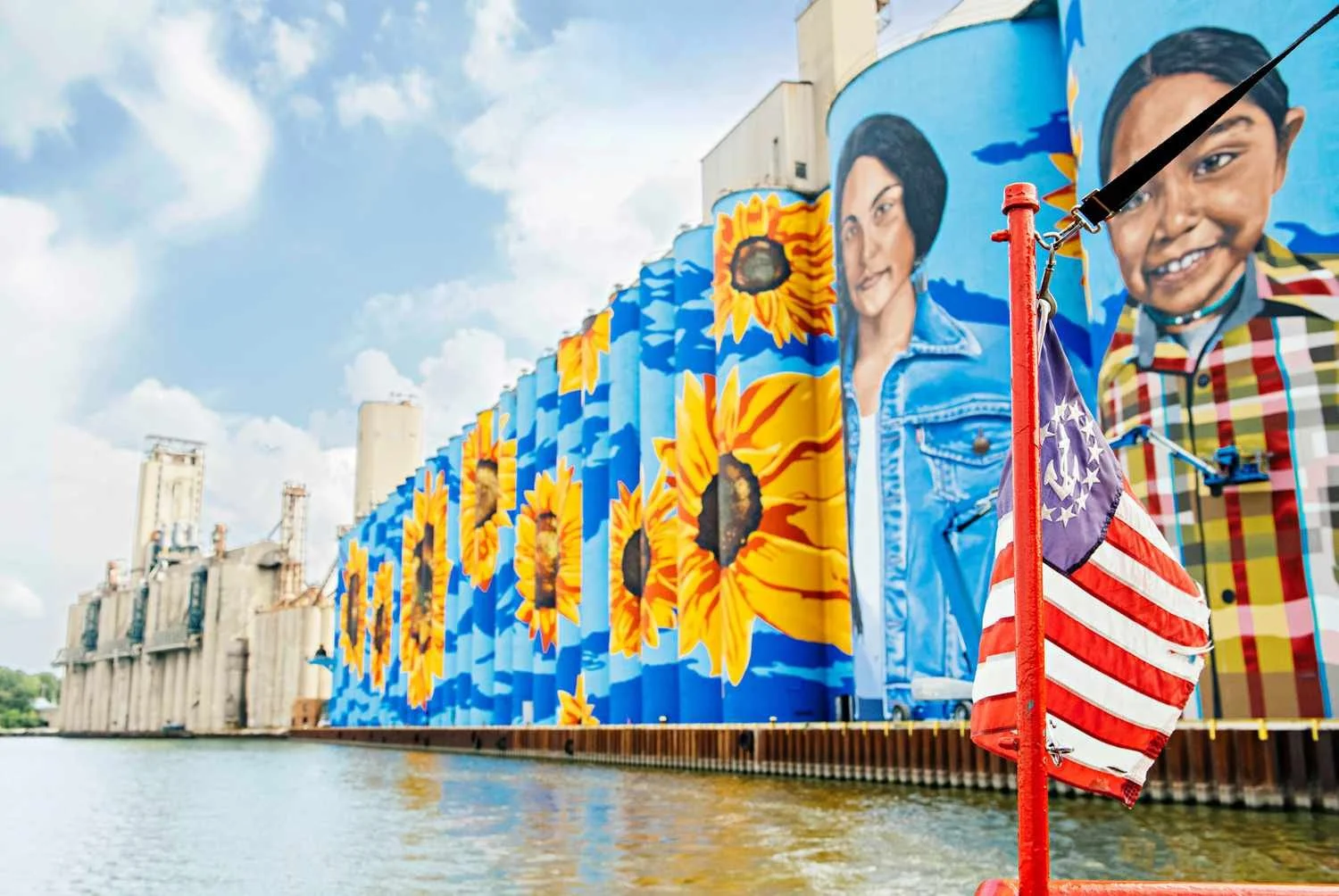 Bright mural with sunflowers and faces painted on industrial silos along a river, with a partly cloudy sky overhead, and a small American flag on a pole in the foreground.