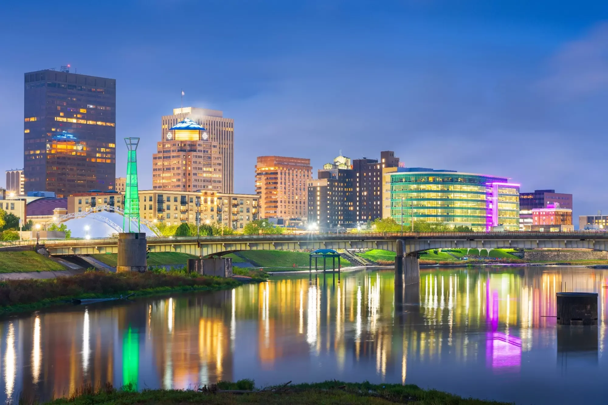Night city skyline reflecting on a river with illuminated buildings, bridges, and colorful lights.